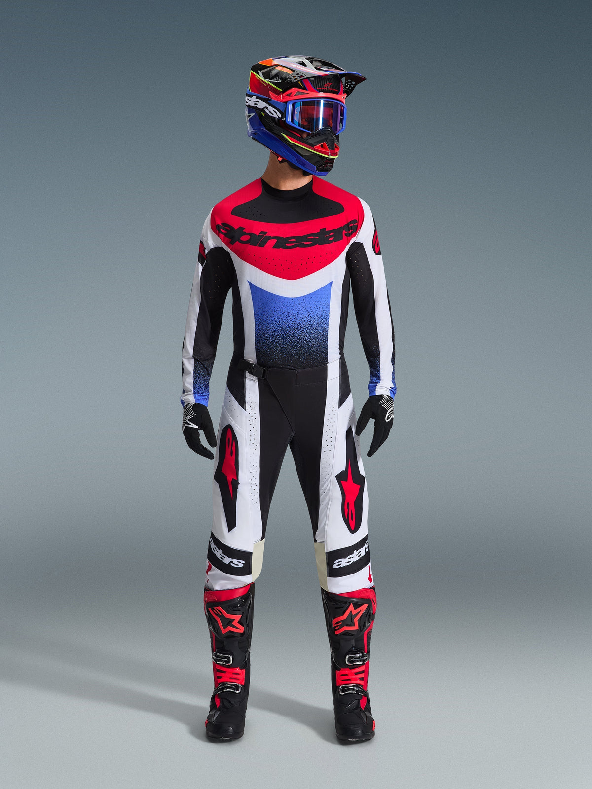Pilote de motocross portant l'ensemble d'équipement Alpinestars Techstar Knif 2026, maillot noir, blanc et rouge avec dégradé bleu et logo, pantalon assorti noir et blanc avec accents rouges, casque multicolore avec lunettes bleues et bottes noires et rouges, tenue de course tout-terrain coordonnée.