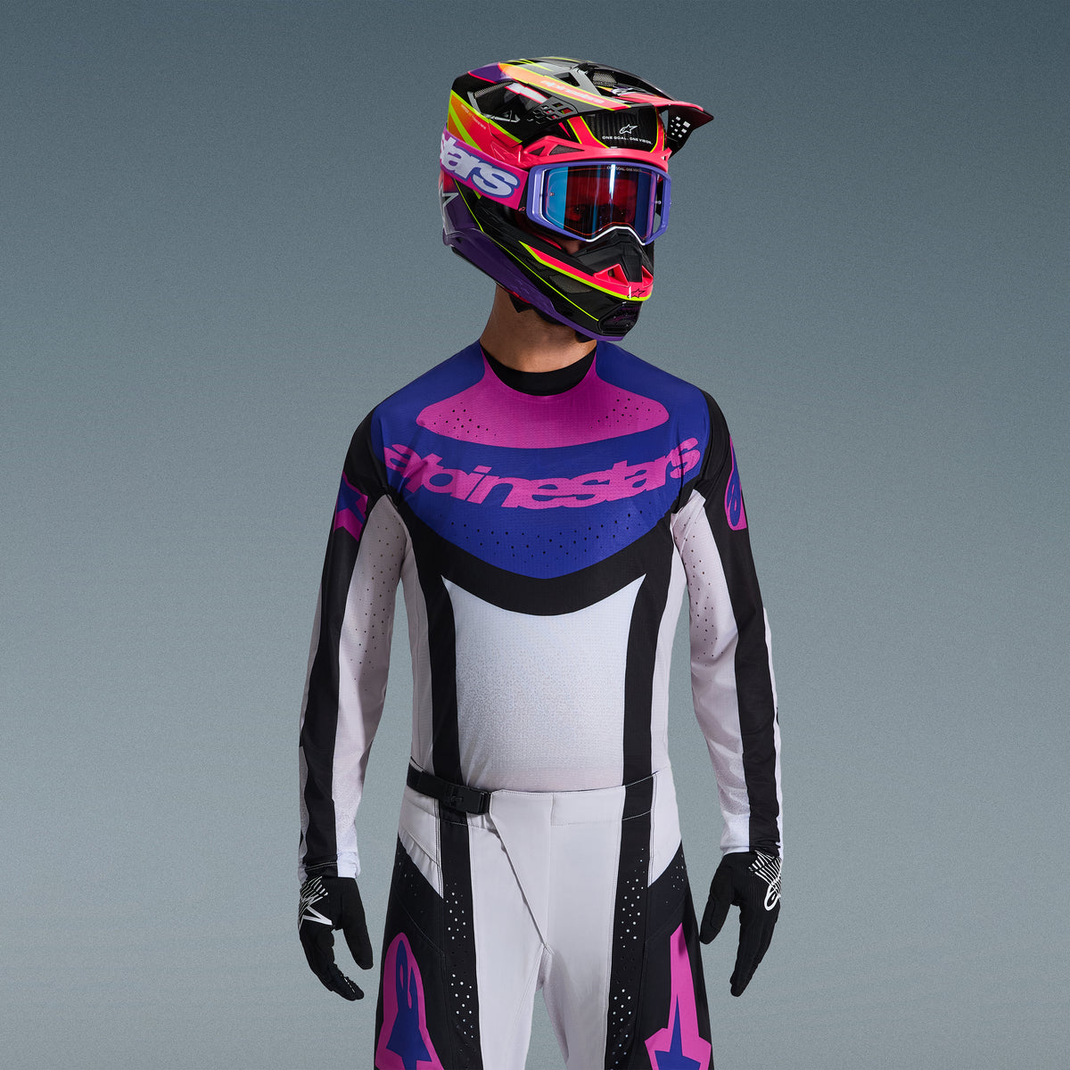Motocross-Fahrer im Alpinestars 2026 Techstar Knif Bekleidungsset, Trikot mit blau-pinker Brustgrafik und weißem Torso, weiße Hose mit schwarzen Einsätzen und pinken Logos, mehrfarbiger Helm in Lila und Pink mit passender Brille, schwarze Handschuhe, professionelle Offroad-Rennausrüstung