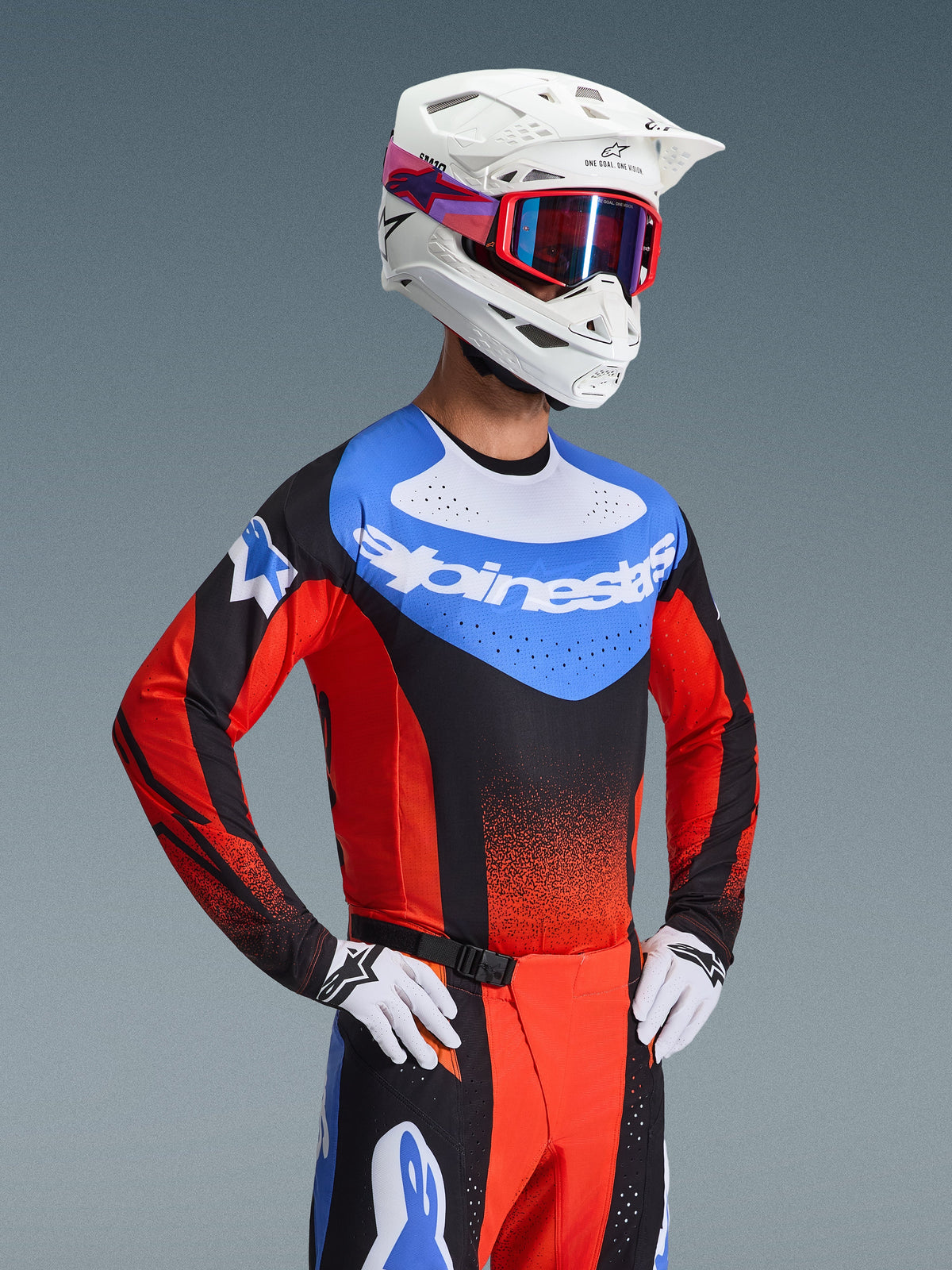 Pilote de motocross portant l'ensemble d'équipement Alpinestars Techstar Knif 2026, maillot bleu avec logo blanc et manches rouges, pantalon assorti noir et rouge avec accents bleus, casque blanc avec lunettes rouges, gants blancs et noirs, tenue de course professionnelle pour motocross
