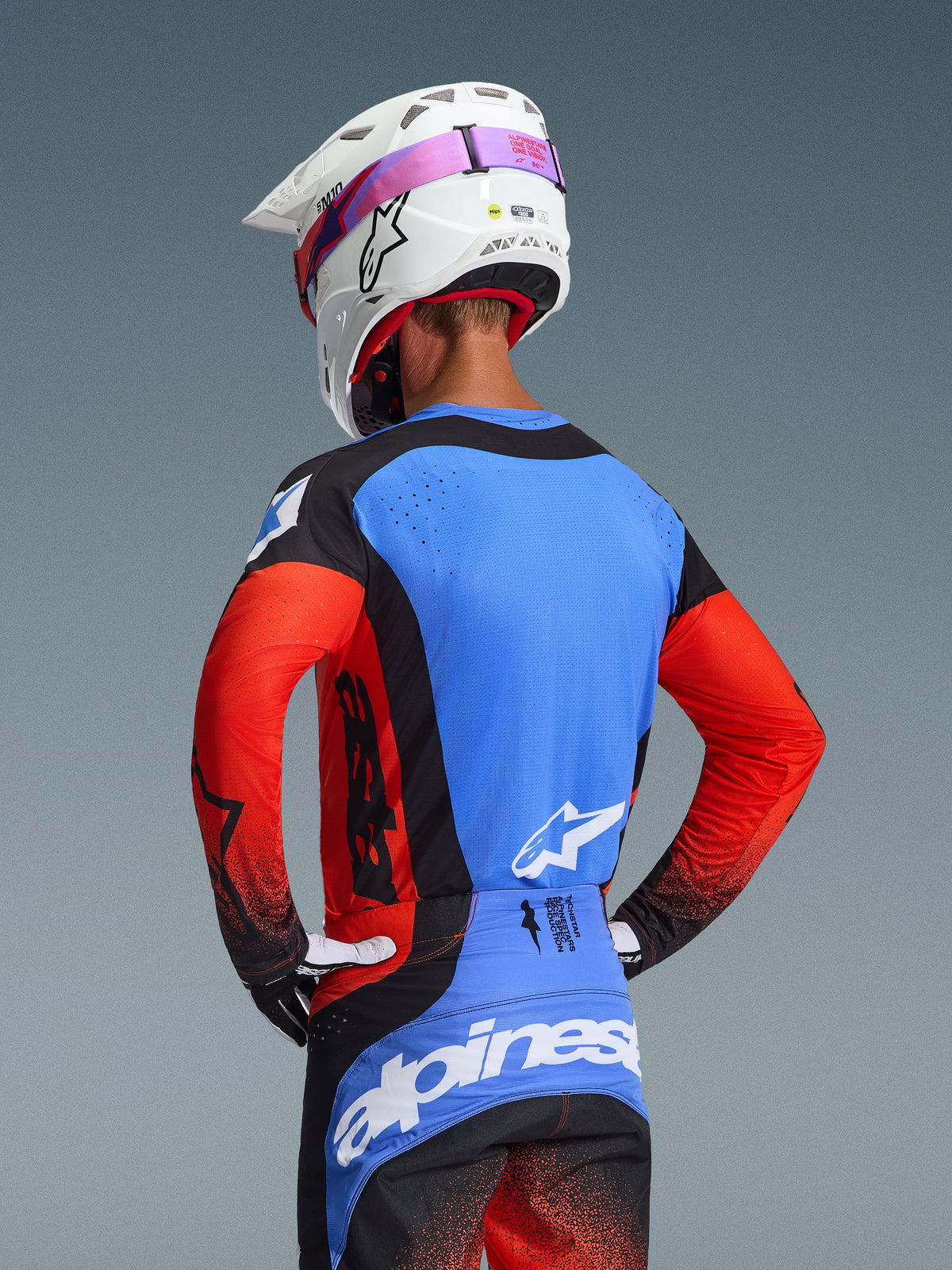 Pilote de motocross vu de dos portant l'ensemble d'équipement Techstar Knif 2026, maillot bleu avec manches rouges et détails noirs, pantalon noir et bleu avec marquage Alpinestars blanc, casque blanc avec sangle de lunettes rose, doté de perforations découpées au laser pour la ventilation