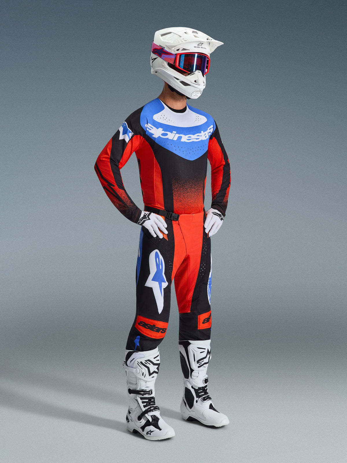 Pilote de motocross portant la tenue Alpinestars 2026 Techstar Knif, le maillot présente un buste bleu avec logo blanc et des manches rouges à dégradés noirs, pantalon noir et rouge assorti avec accents d'étoiles bleues, casque blanc avec lunettes rouges et bleues, bottes blanches et noires, et gants blancs.