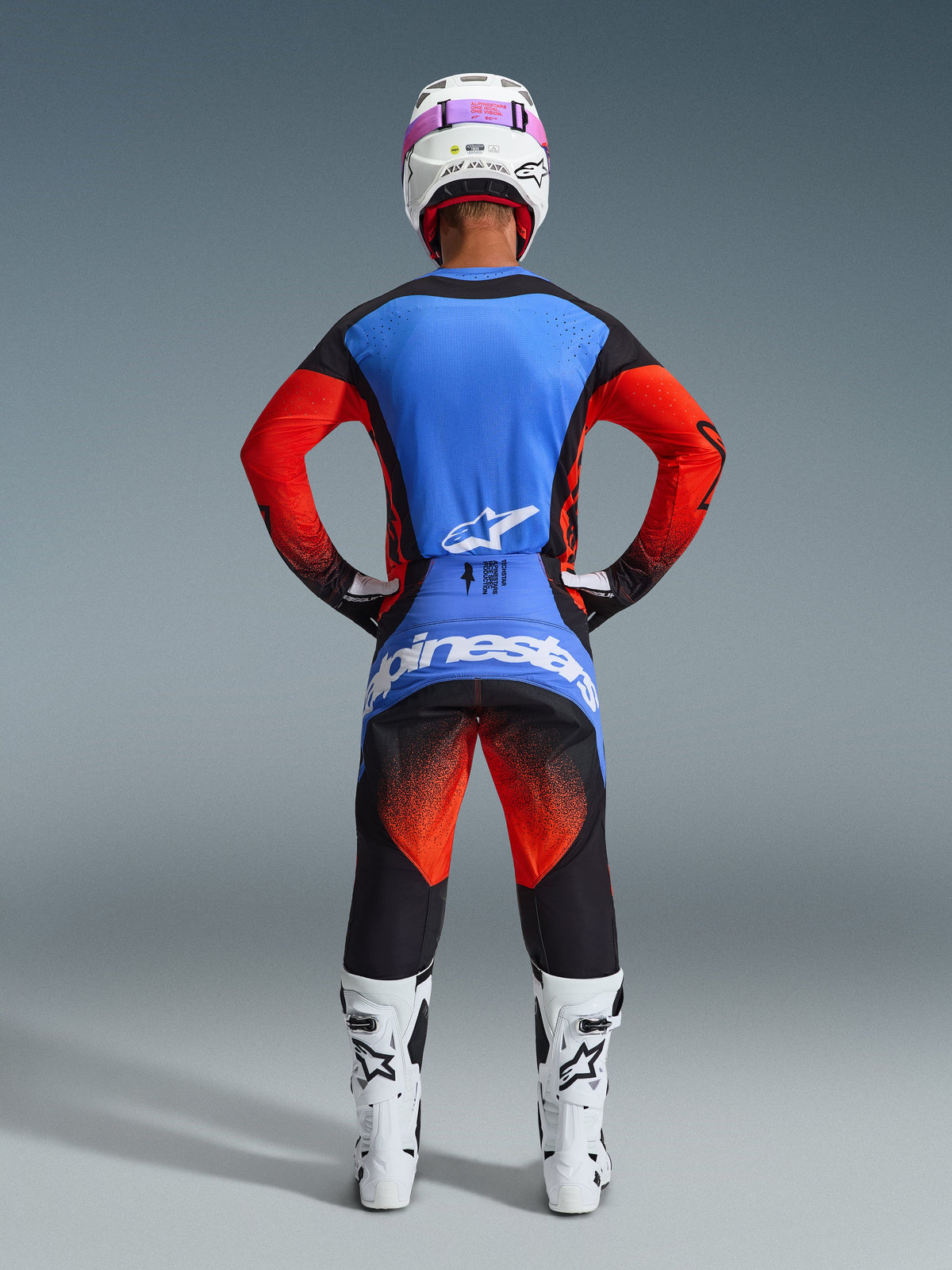 Vue arrière d'un pilote de motocross portant l'ensemble Alpinestars Techstar Knif 2026, maillot bleu et rouge avec accents noirs, pantalon noir avec dégradé rouge sur les cuisses et logo bleu, avec bottes de course blanches et casque blanc avec lunettes roses.
