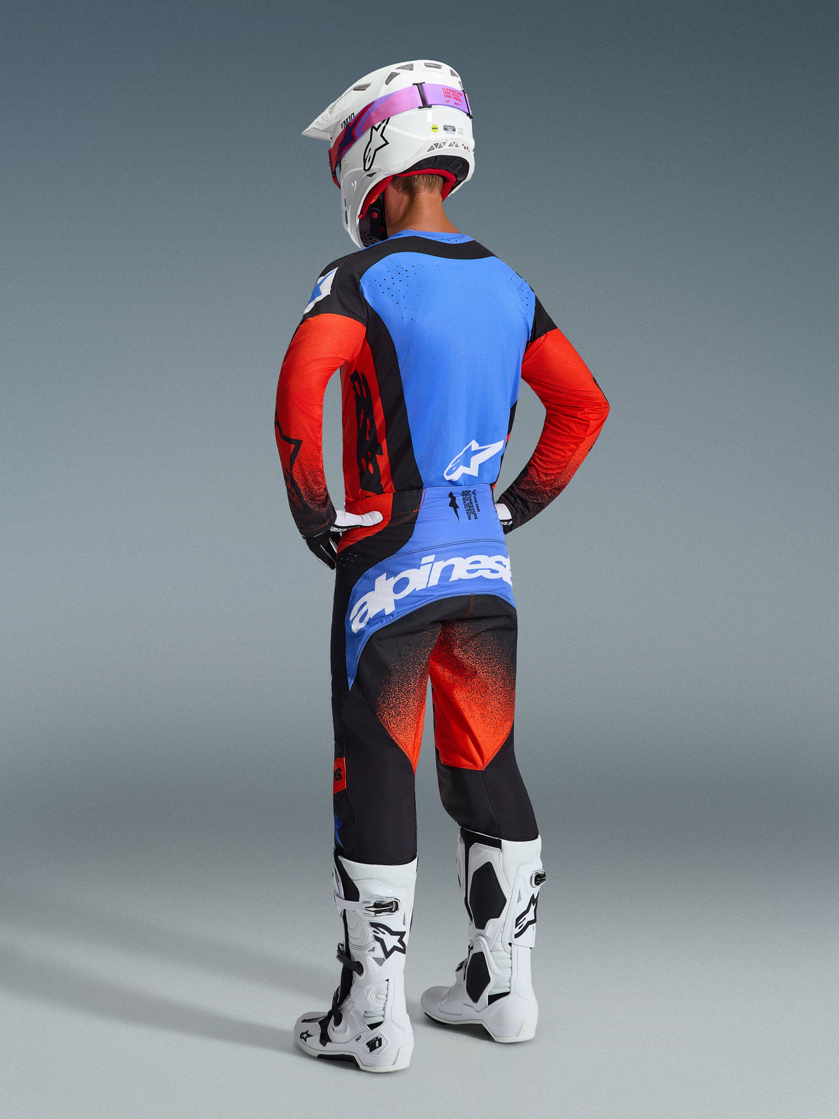 Vue arrière d'un pilote de motocross portant l'ensemble Alpinestars Techstar Knif 2026, maillot bleu et rouge avec accents noirs, pantalon noir et rouge assorti avec un grand logo bleu sur l'assise, casque blanc et bottes de motocross blanches, équipement professionnel de course tout-terrain