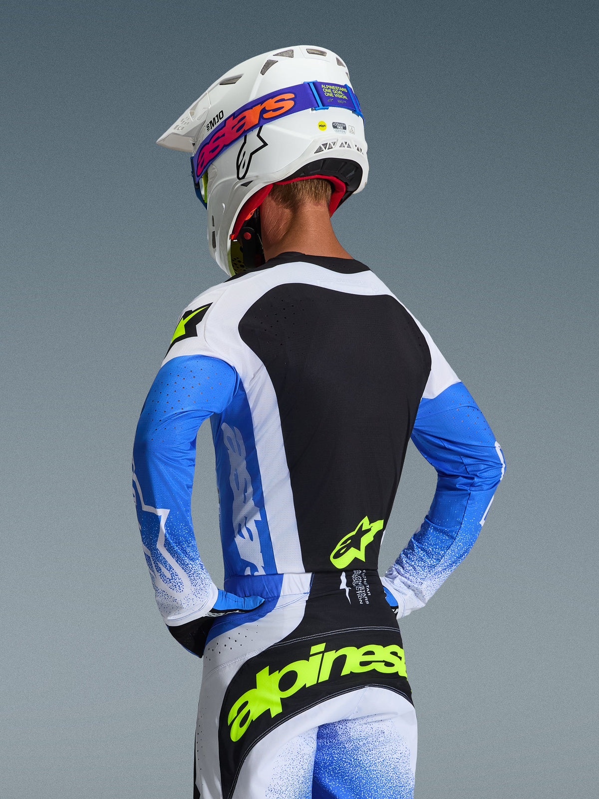 Rückansicht eines Motocross-Fahrers im Alpinestars 2026 Techstar Knif Jersey in Ucla Blue, Schwarz und Fluogelb, kombiniert mit der passenden Hose und einem weißen Supertech M10 Helm, athletische Rennpassform für Offroad-Performance.