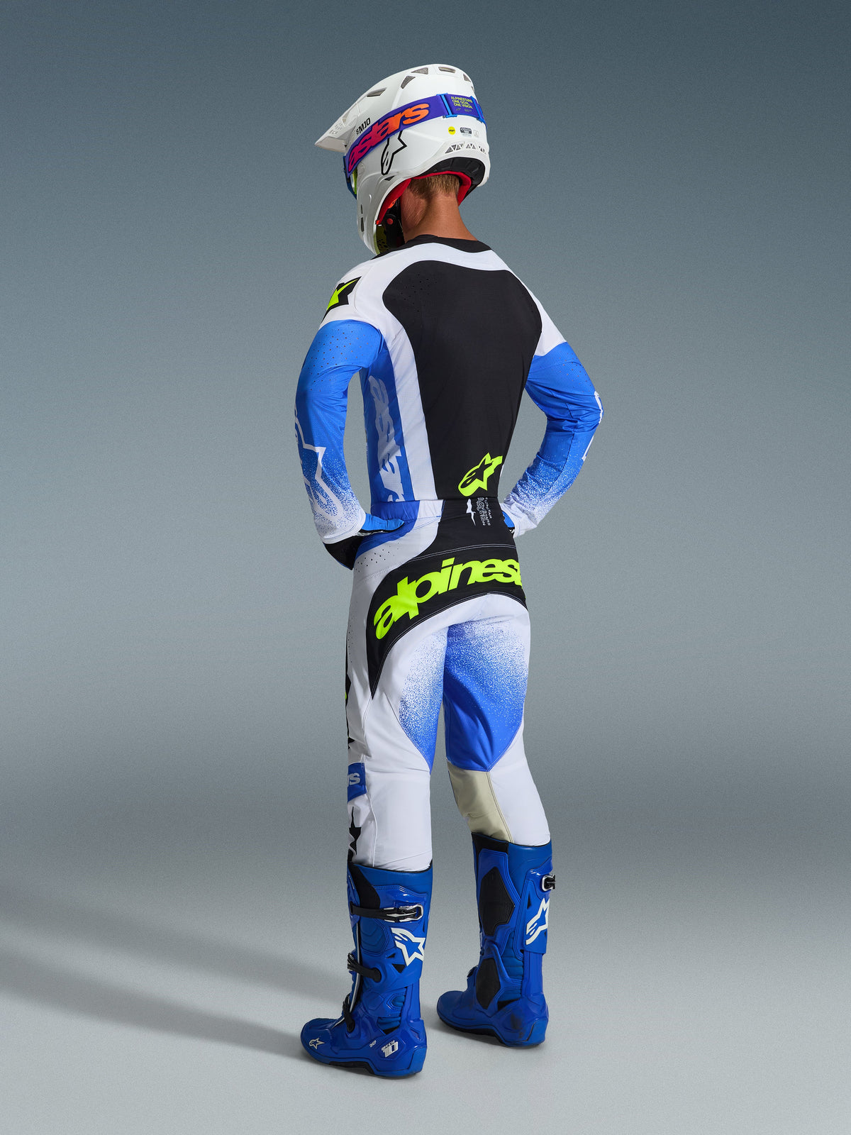 Motocross-Fahrer von hinten gesehen, trägt das komplette Alpinestars 2026 Techstar Knif Gear Set in Ucla Blue und Schwarz, mit einem Jersey mit schwarzem Rückeneinsatz und neongelbem Logo, einer dazu passenden blau-weißen Hose mit markantem gelbem Branding, blauen Motocross-Stiefeln und einem weißen Rennhelm.