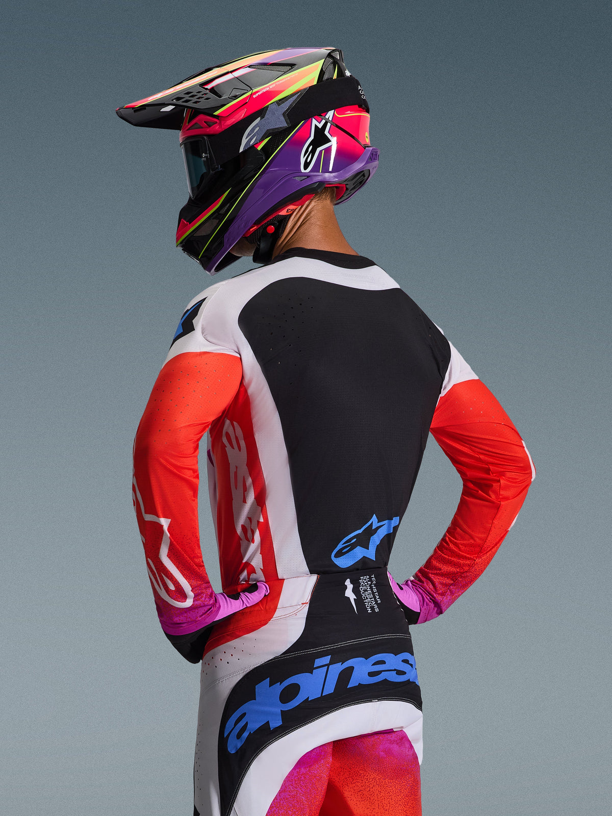 Pilote de motocross de dos portant le maillot Alpinestars 2026 Techstar Knif en coloris Gris clair Orange Fluo Noir, doté d'un panneau dorsal noir avec ventilation découpée au laser, de manches orange et de panneaux latéraux gris clair, coordonné à un pantalon assorti et un casque coloré.