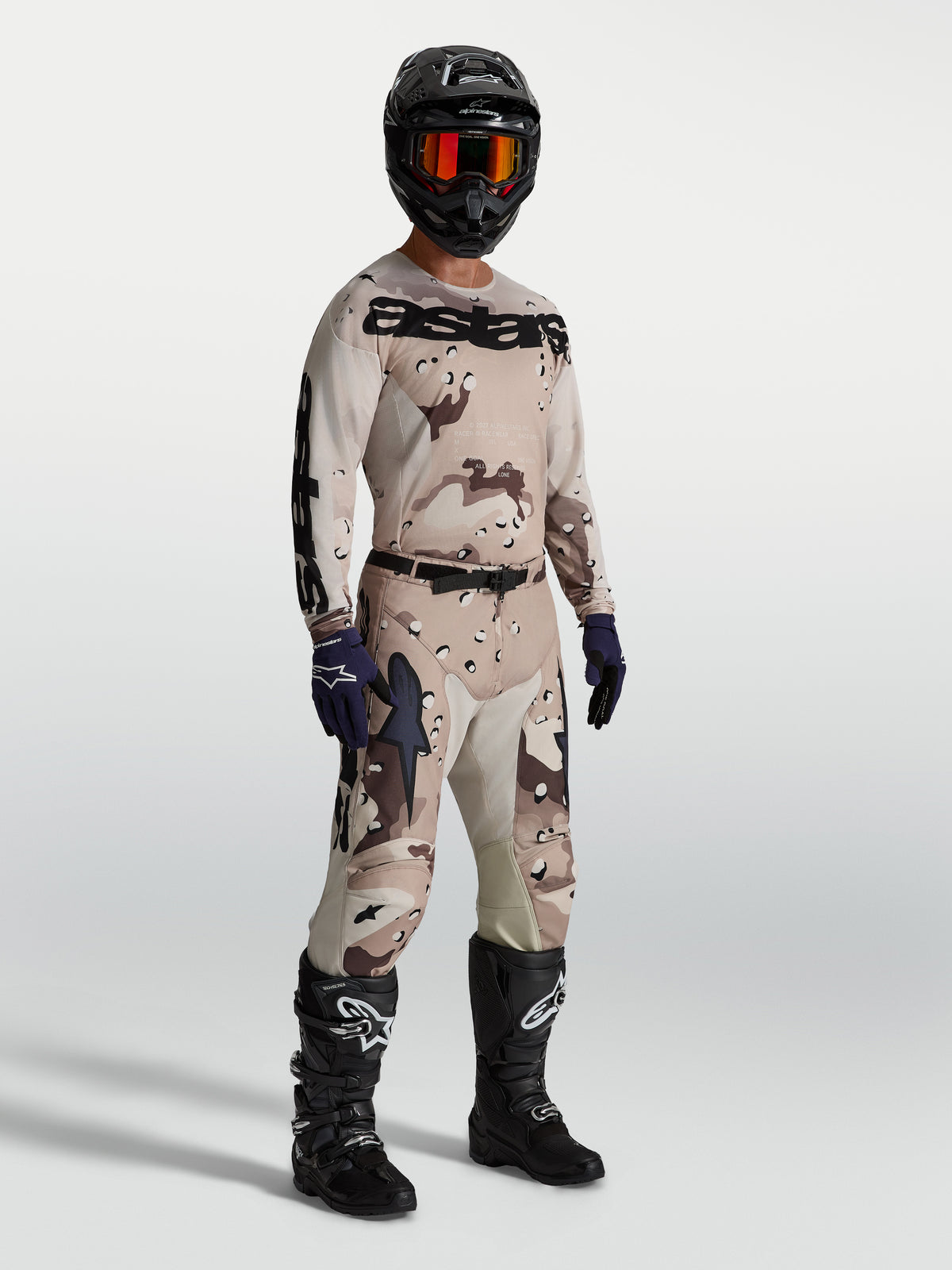 Alpinestars Racer Lone Jersey, heren-motocrossjersey in desert camo en zwart, vochtafvoerende lichtgewicht constructie, gedragen door een model met bijpassende broek en zwarte motorlaarzen.