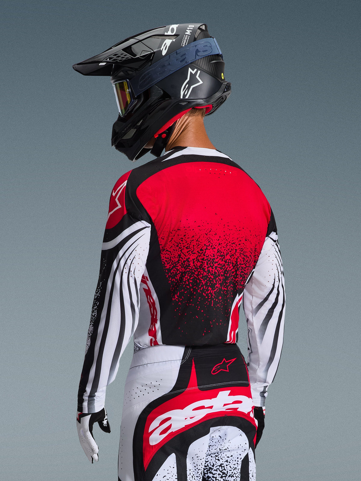 Motocross-Fahrer von hinten im 2026 Techstar Nomur Trikot, rote Rückseite mit schwarzem Farbverlauf, weiße Ärmel mit schwarzen Akzenten, getragen mit passender rot-weißer Hose, schwarzem Helm und weißen Handschuhen, professionelle Offroad-Rennausrüstung.