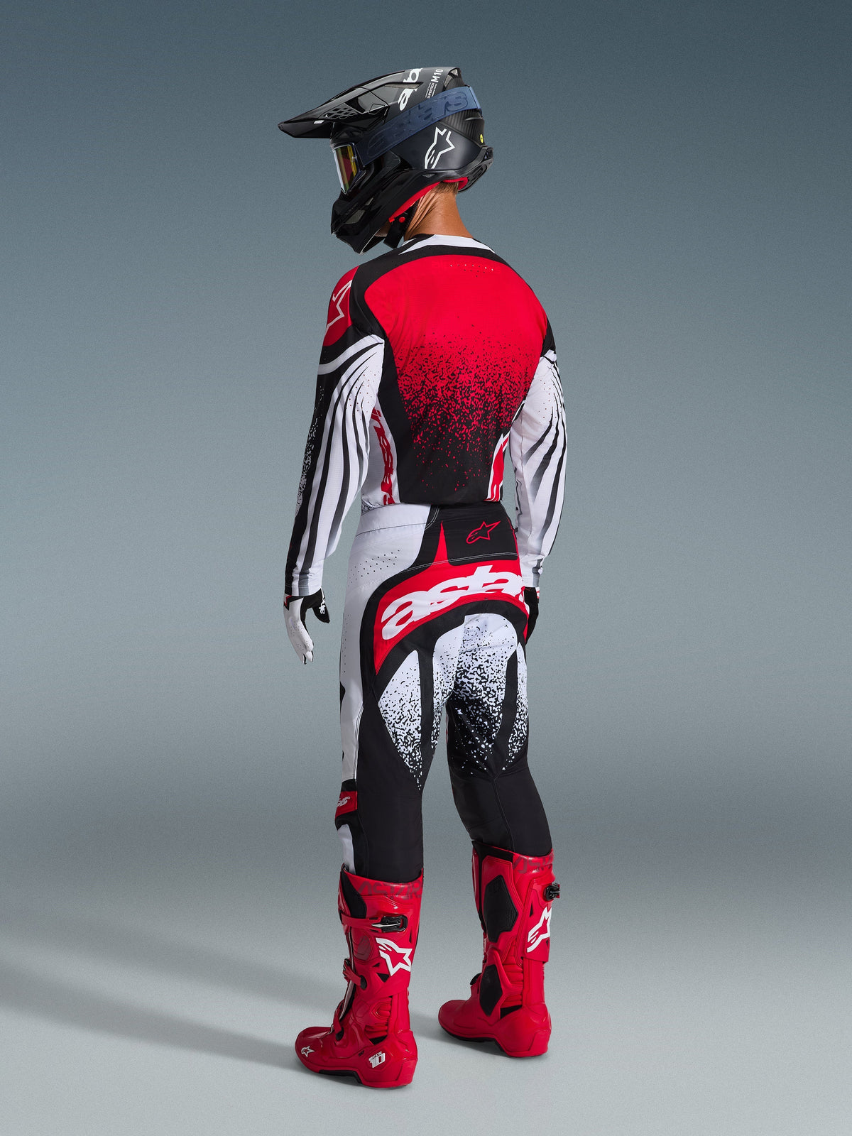 Rückansicht eines Motocross-Fahrers im Alpinestars 2026 Techstar Nomur Trikot in Schwarz-Weiß-Rot, mit rotem Farbverlauf am Rücken und wellenförmigen Ärmelmustern, kombiniert mit passender Motocross-Hose, roten Stiefeln und schwarzem Helm für eine abgestimmte Offroad-Rennausrüstung.