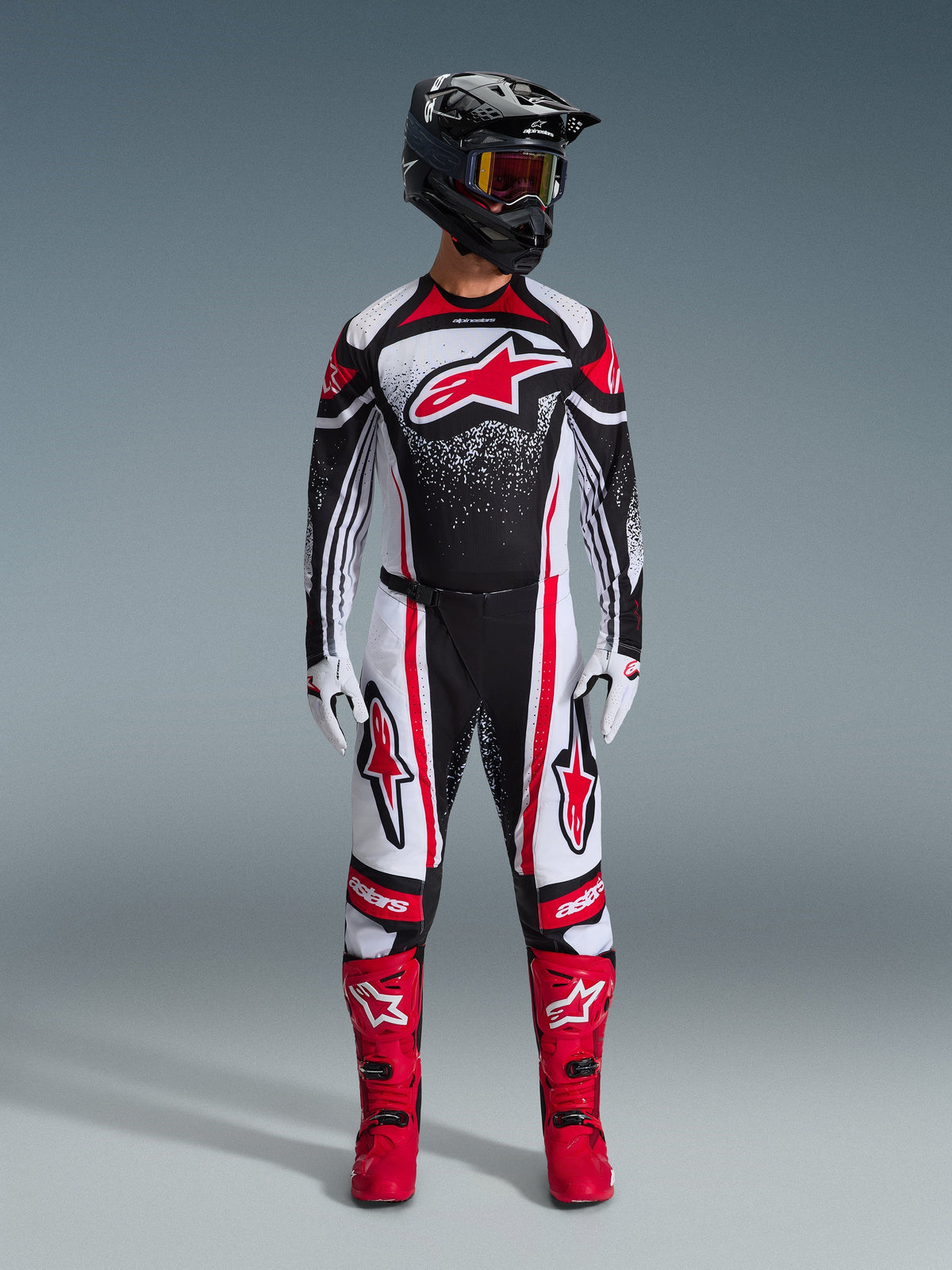 Motocross-Fahrer in kompletter Alpinestars Rennausrüstung, 2026 Techstar Nomur Trikot in Schwarz-Weiß-Rot, passende Hose mit roten Akzenten, schwarzer Helm und leuchtend rote Stiefel, großes rotes Astars-Logo auf der Brust mit gesprenkeltem Verlaufsmuster, abgestimmtes Outfit für den Offroad-Wettbewerb.