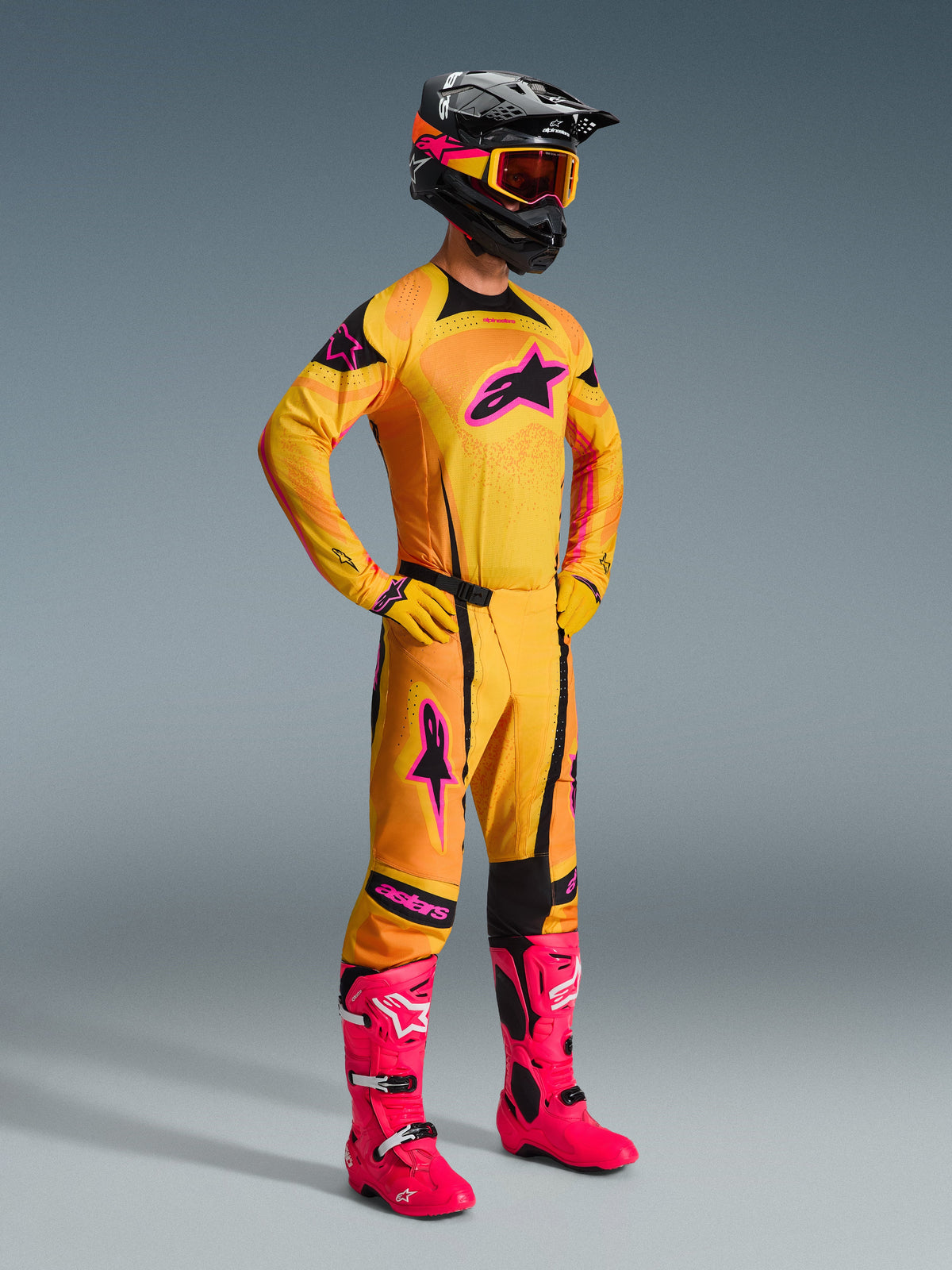 Motocross-Fahrer im Alpinestars 2026 Techstar Nomur Ausrüstungsset, Trikot und passende Hose in Orange, Gelb und Pink mit schwarzen Akzenten, mit einem großen pinkfarbenen Stern-Logo auf der Brust, leuchtend pinkfarbene Rennstiefel, schwarzer Helm mit orange- und pinkfarbenen Grafiken und gelbe Goggles, professionelle Offroad-Wettkampfausrüstung.