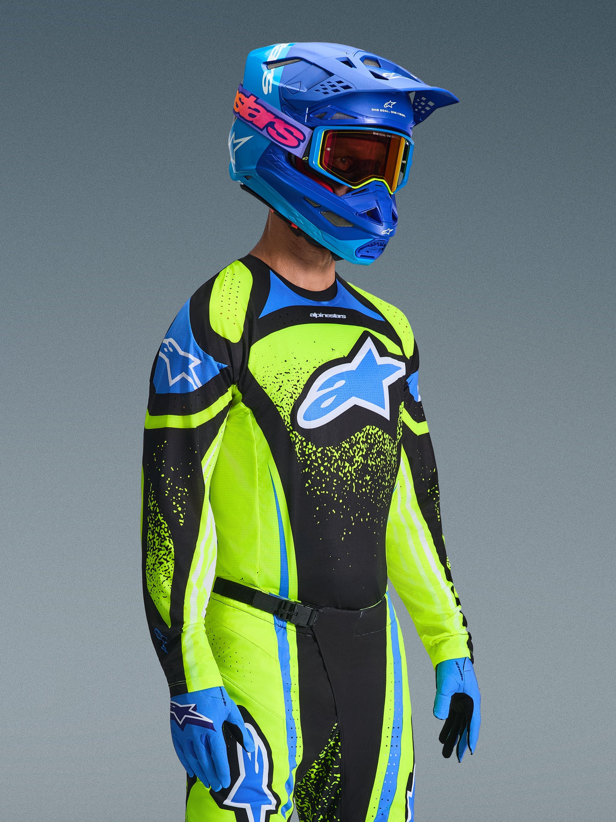Pilote de motocross portant la tenue Alpinestars 2026 Techstar Nomur, maillot jaune fluo et bleu avec panneaux noirs, grand logo bleu et blanc sur la poitrine, casque bleu assorti avec lunettes orange, gants bleus et noirs, équipement haute performance coordonné pour la course tout-terrain