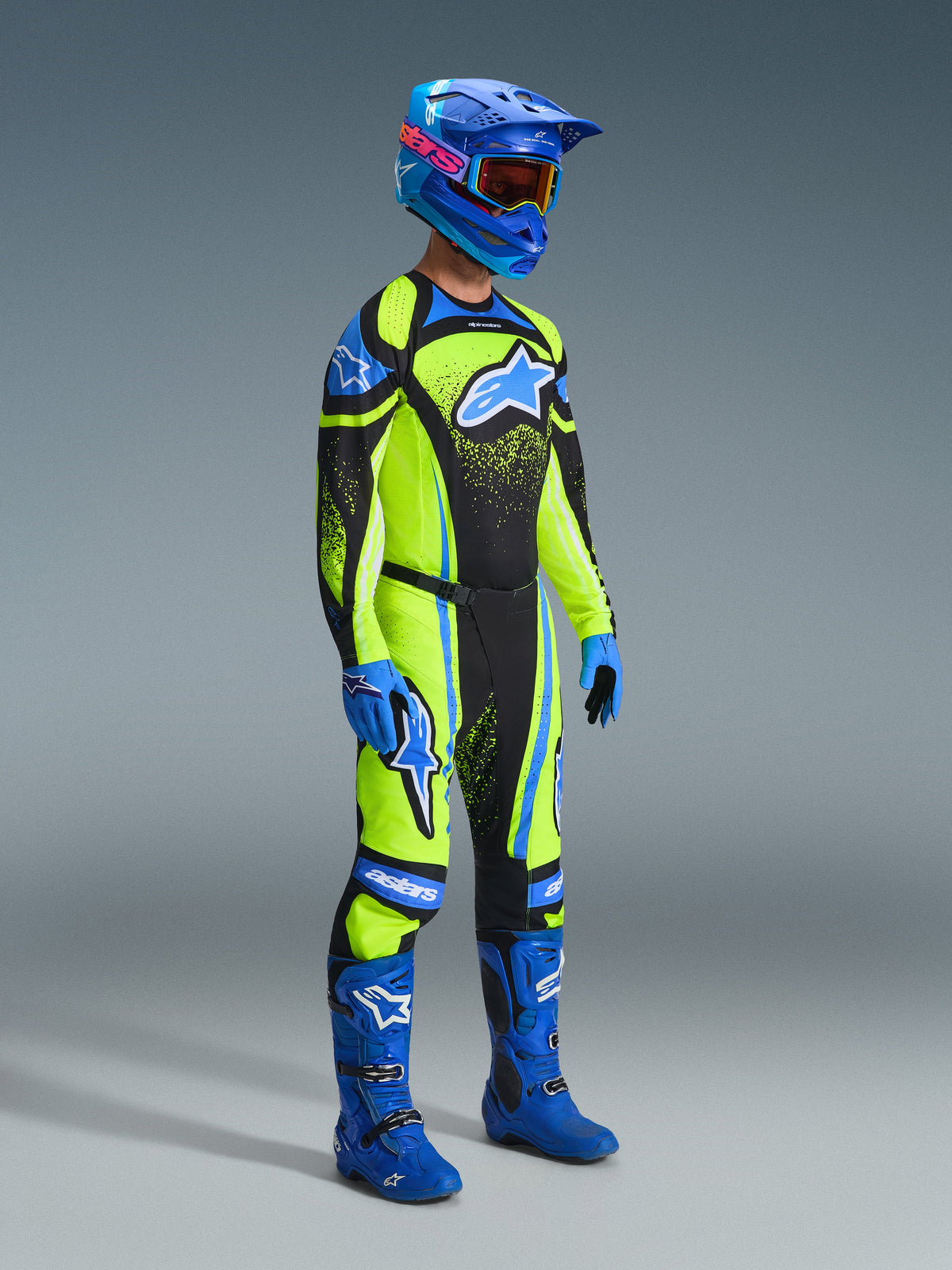 Pilote de motocross portant l'ensemble Alpinestars Techstar Nomur 2026, maillot et pantalon vert citron éclatant avec empiècements noirs et détails bleus, grand logo étoile bleu sur la poitrine et les cuisses, casque bleu avec lunettes orange, bottes et gants bleus, tenue de course complète pour motocross