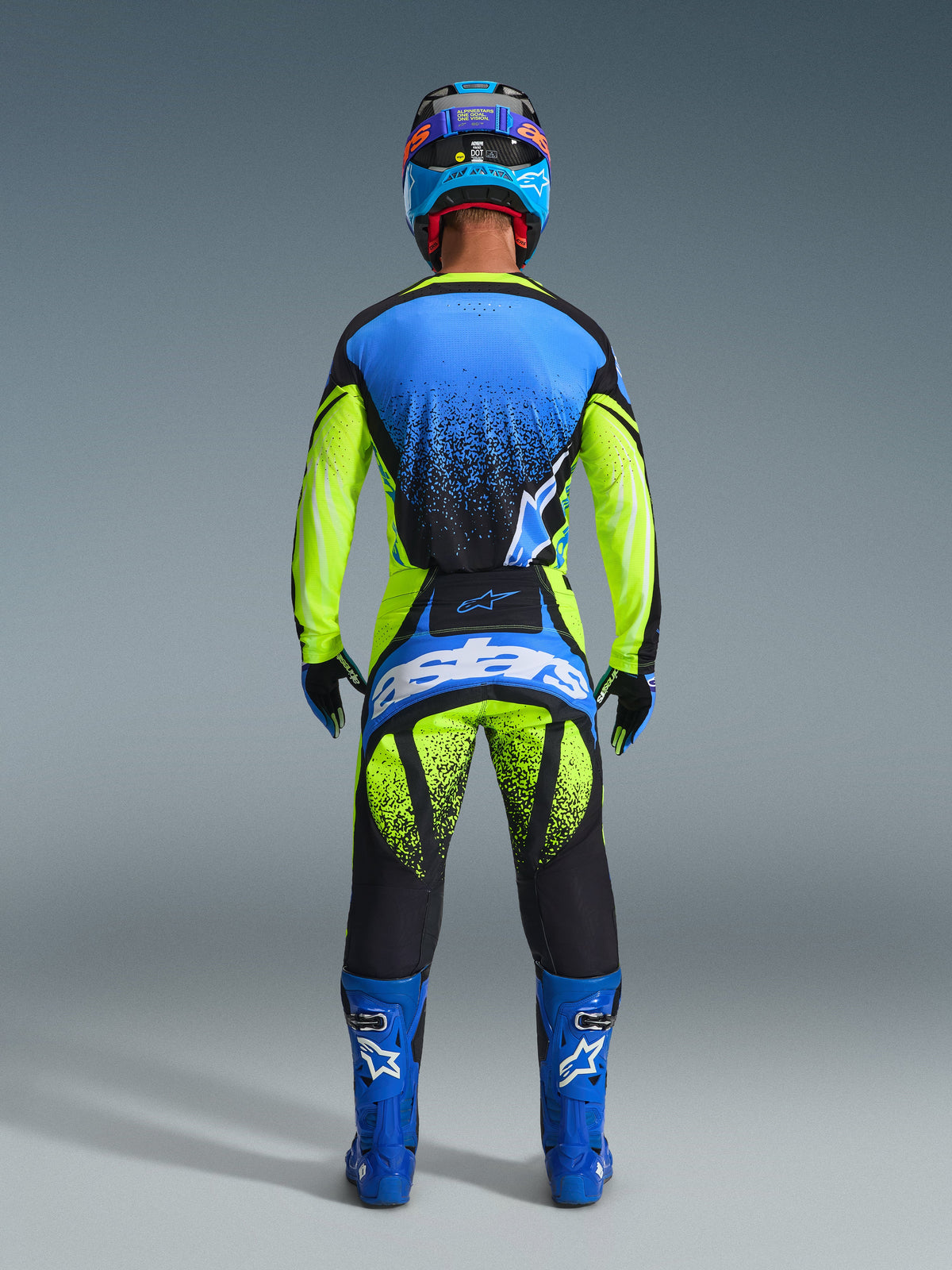 Vue arrière d'un pilote de motocross portant l'ensemble Alpinestars 2026 Techstar Nomur. La tenue comprend un maillot bleu et jaune fluo avec des accents noirs à motif de pulvérisation, un pantalon assorti jaune fluo et noir avec un grand logo « astars » bleu sur l'assise, des bottes bleues coordonnées avec logos blancs et un casque bleu et noir. Équipement de course professionnel complet conçu pour la performance et la visibilité.