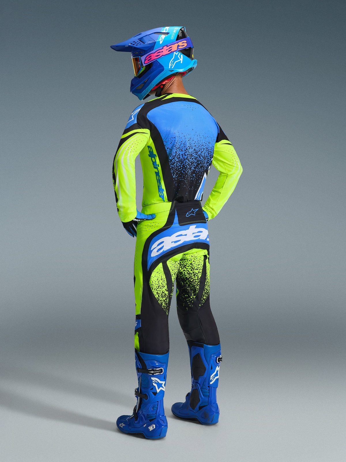 Pilote de motocross portant la tenue Alpinestars Techstar Nomur 2026, maillot jaune fluo et bleu marine avec touches de bleu, pantalon assorti jaune et noir avec large logo 'astars', casque bleu aux accents roses et lunettes bleues, bottes bleu vif, équipement de motocross haute performance coordonné
