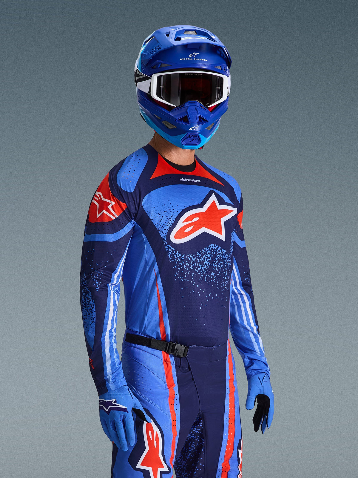 Pilote de motocross portant le maillot Alpinestars Techstar Nomur 2026, tenue de course bleu marine foncé, orange et bleu UCLA, grand logo Astars orange, casque et gants bleus assortis, tenue de compétition tout-terrain athlétique.
