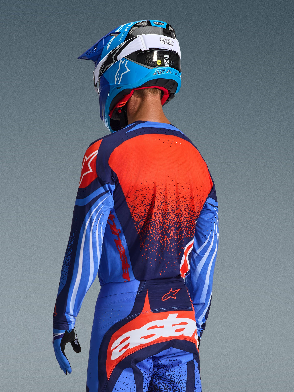 Pilote de motocross vu de dos portant le maillot Alpinestars Techstar Nomur 2026, bleu marine avec détails orange et bleu UCLA, grand graphisme texturé orange au dos, pantalon assorti et casque de course bleu, tenue professionnelle pour le tout-terrain.