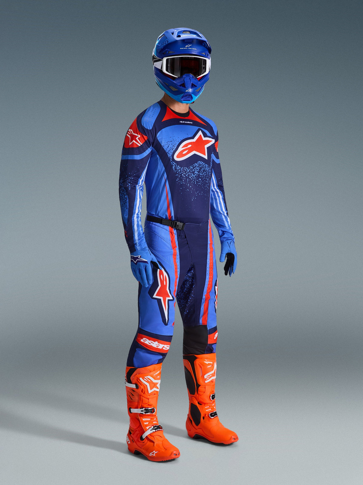 Pilote de motocross portant un équipement de course complet, maillot Techstar Nomur bleu et bleu marine avec logo Alpinestars orange, pantalon assorti avec accents orange, casque bleu avec lunettes blanches, bottes orange vif et gants bleus, tenue coordonnée pour la course tout-terrain