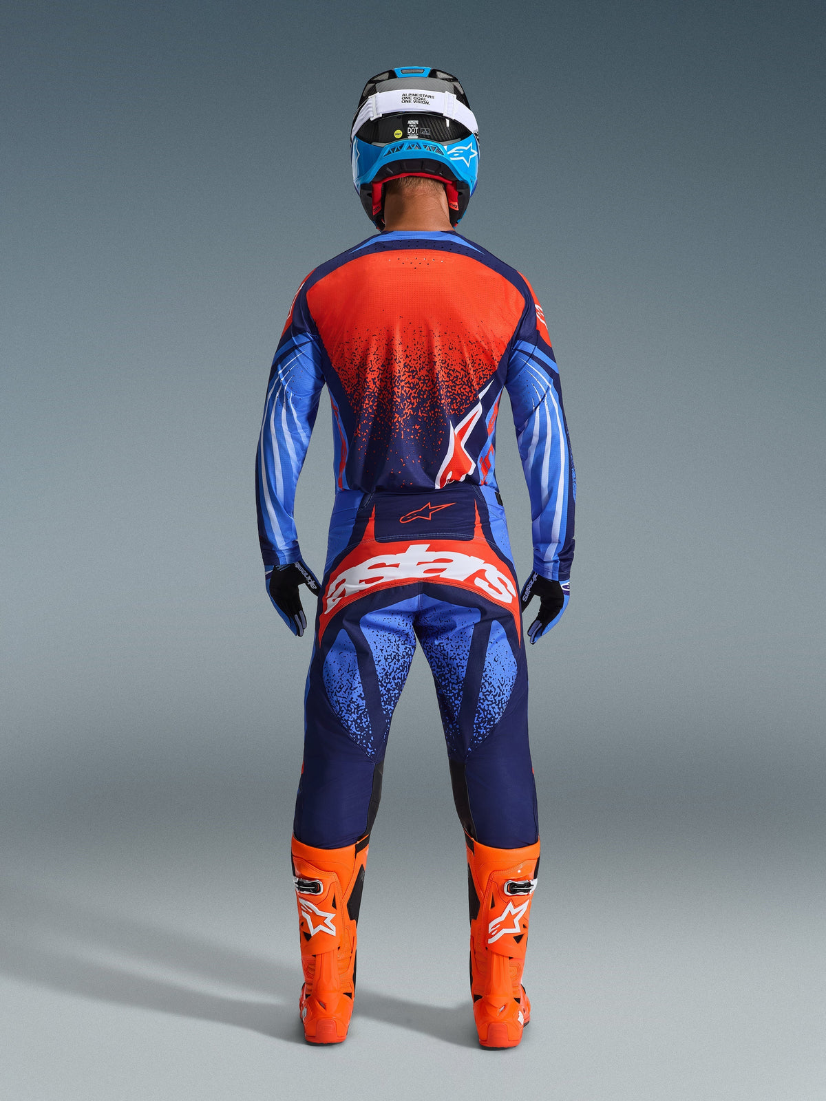 Pilote de motocross de dos portant le maillot Techstar Nomur 2026 Alpinestars et le pantalon assorti, coloris bleu marine foncé et orange avec accents bleu UCLA, bottes orange vif, casque bleu et blanc et gants noirs, équipement complet de course tout-terrain professionnel