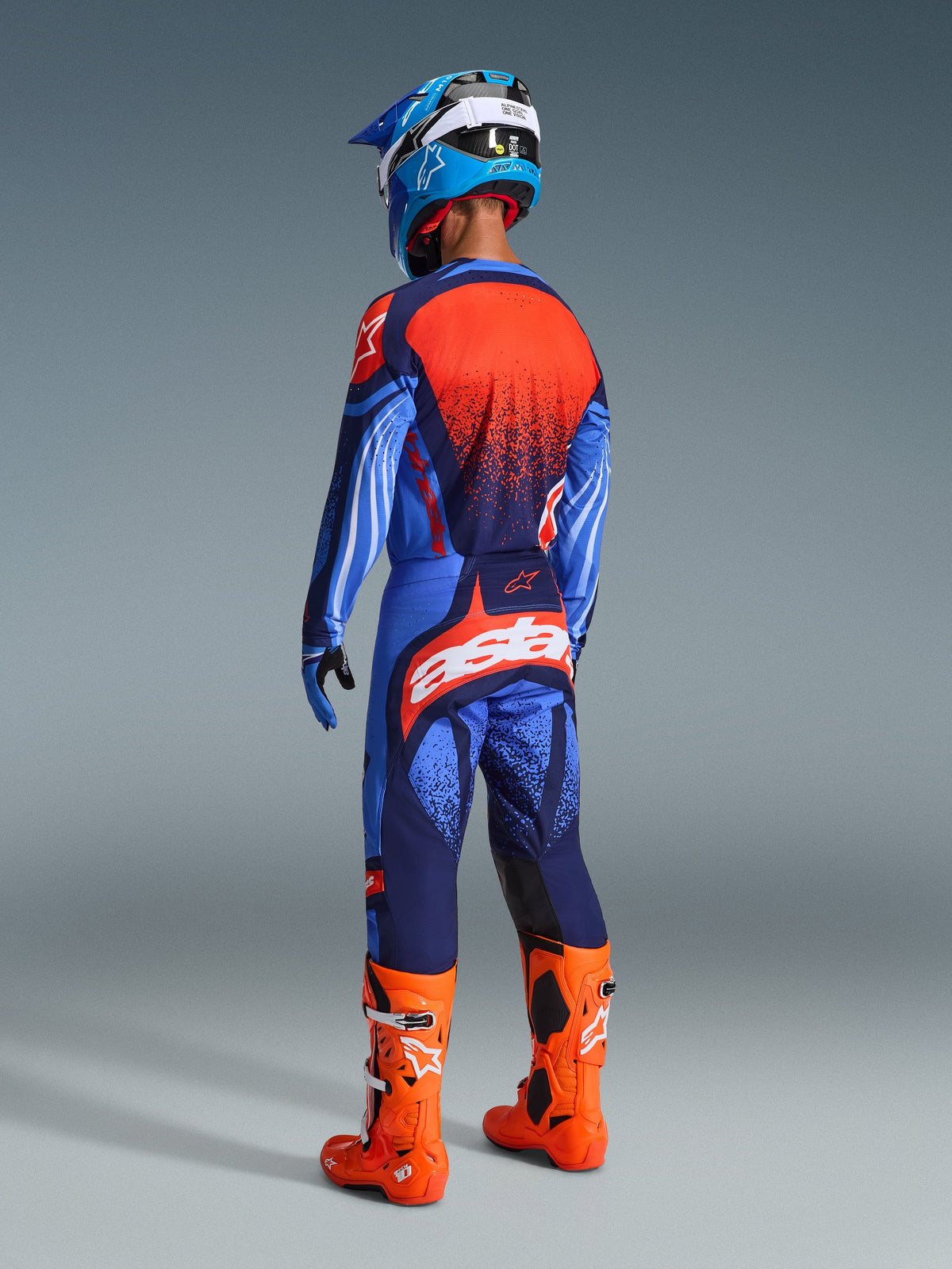 Motocrossrijder vanaf de achterkant gezien in de 2026 Techstar Nomur Gear Set, donkerblauw en UCLA-blauw shirt met oranje paneel, bijpassende broek met oranje 'astars'-logo, feloranje motorcrosslaarzen en een blauw-witte helm, gecoördineerde competitie-race-uitrusting