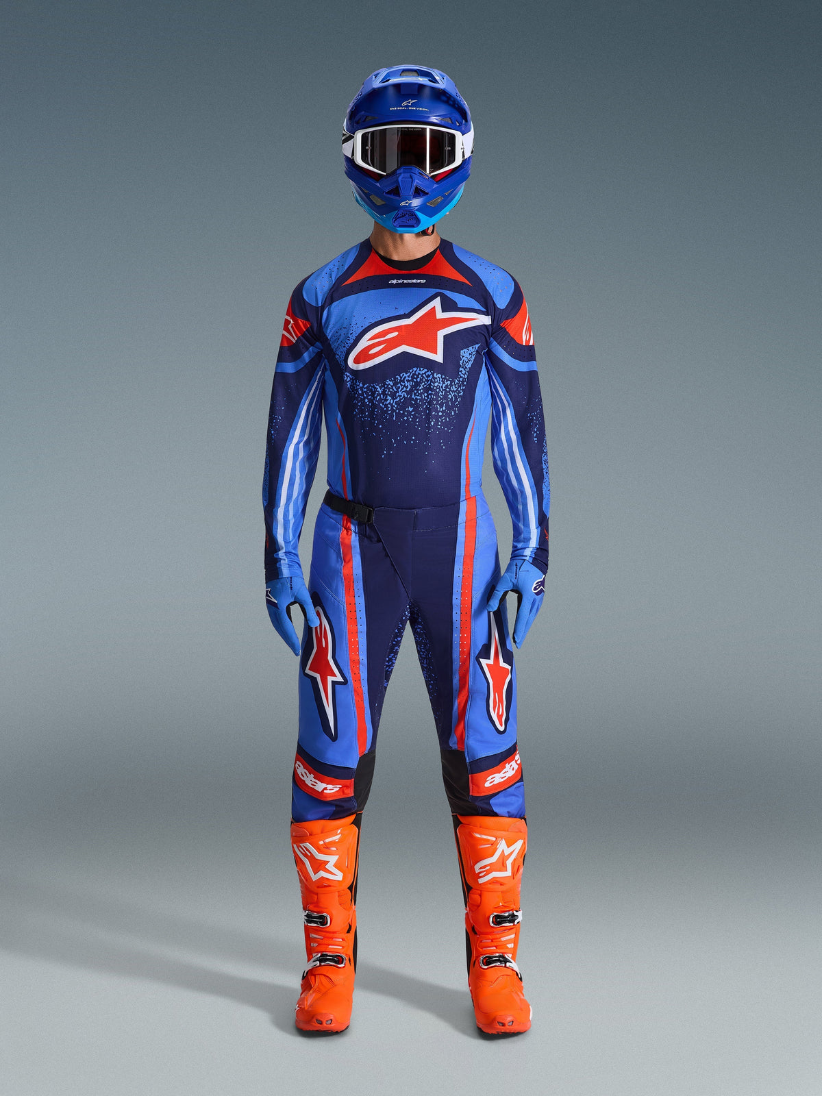 Pilote de motocross portant le maillot Alpinestars Techstar Nomur 2026 en bleu marine foncé, orange et bleu UCLA, associé à un pantalon de motocross assorti, un casque bleu avec lunettes et des bottes de course orange vif, tenue de compétition tout-terrain complète et coordonnée