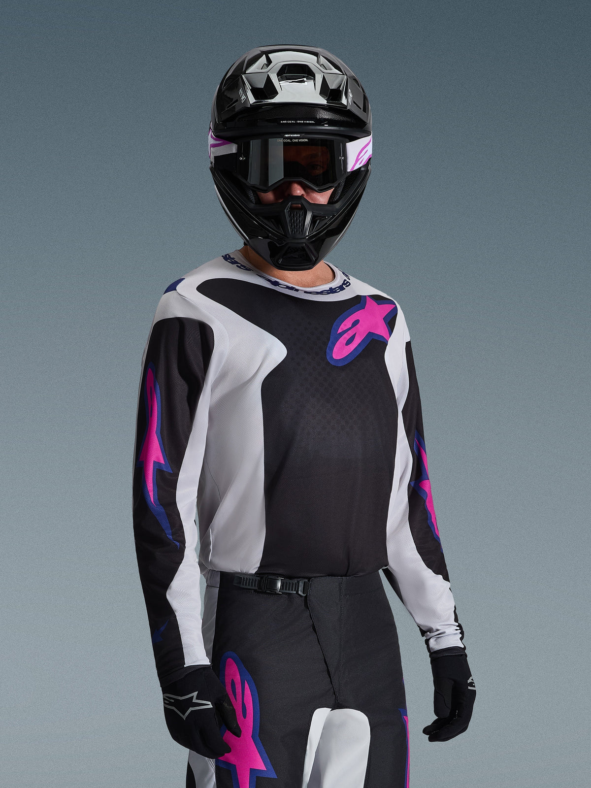 Motorcrossrijder die de Alpinestars 2026 Fluid Grid Jersey draagt, zwart, lichtgrijs en paars, voorzien van grote roze en paarse sterlogo's, gecombineerd met een bijpassende broek, zwarte helm en bril, complete off-road race-uitrusting