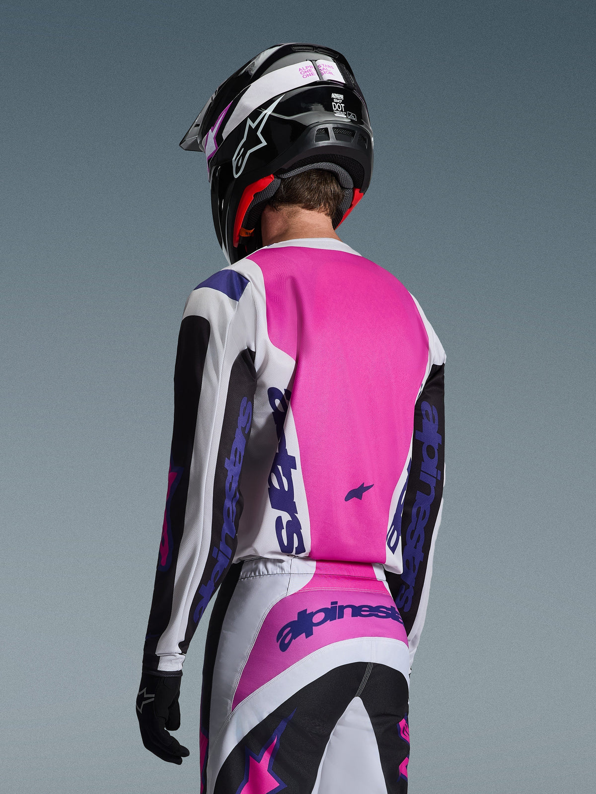 Motocrossrijder van achteren gezien in de Alpinestars 2026 Fluid Grid Jersey in zwart, wit en paars, bijpassende broek met opvallende branding, zwarte helm met roze accenten en zwarte handschoenen, complete gecoördineerde uitrusting voor off-road prestaties.