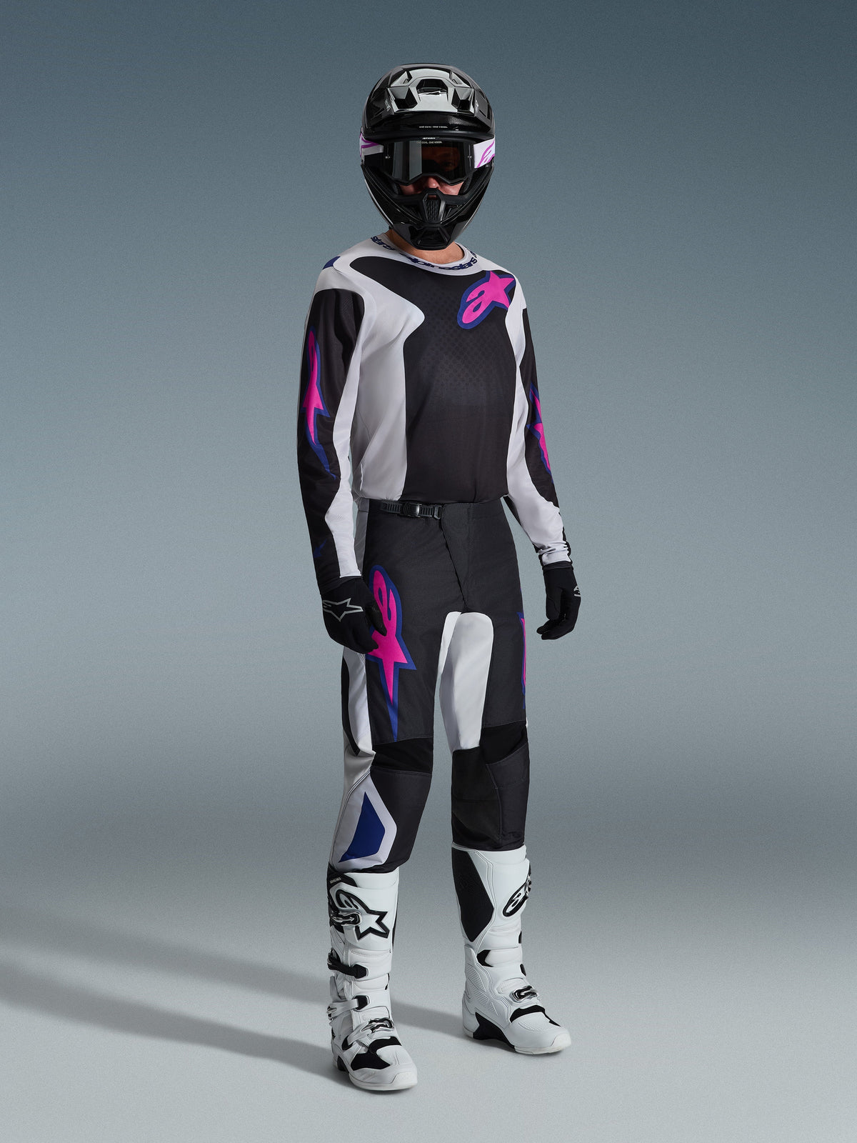 Motorcrossrijder in volledige race-uitrusting, 2026 Fluid Grid Jersey in zwart met lichtgrijze en paarse accenten, bijpassende Fluid Grid Pants, zwarte helm met gecoördineerde bril, wit-zwarte laarzen en zwarte handschoenen, professionele studio-opstelling voor off-road racen.