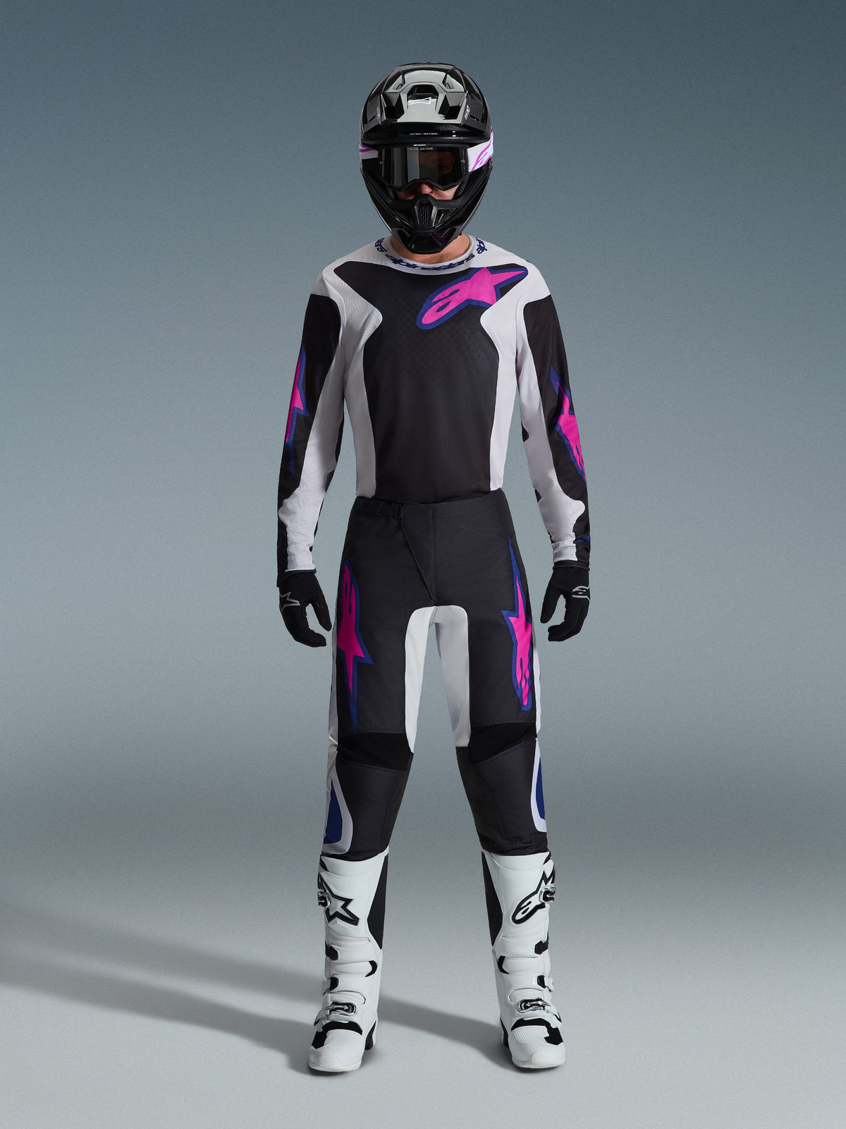Alpinestars motorcrosser in volledige race-uitrusting, 2026 Fluid Grid Jersey en bijpassende broek in zwart en lichtgrijs met levendige paarse accenten, zwarte helm met paarse bril, witte motorcrosslaarzen en zwarte handschoenen, gecoördineerde outfit voor off-road prestaties