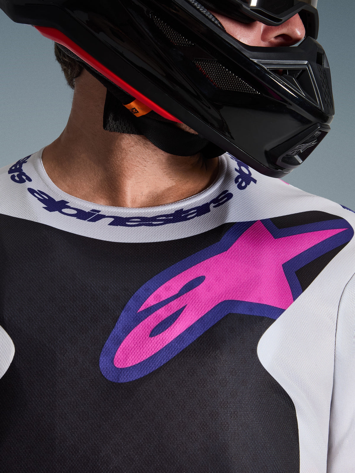 Alpinestars 2026 Fluid Grid Jersey, motocross shirt, zwart, lichtgrijs en paars, close-up van de borst en hals met groot roze sterlogo, sportkraag met merknaam, gedragen met een zwarte helm, hoogwaardige off-road uitrusting