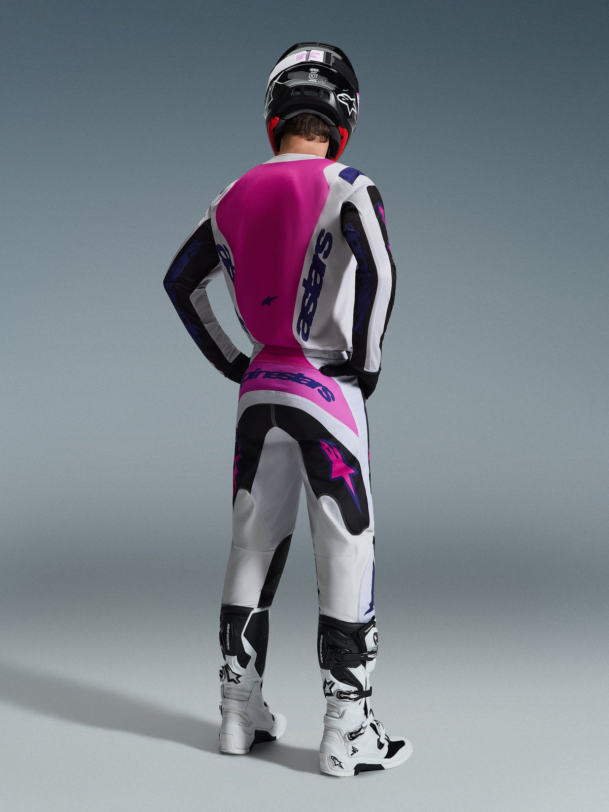 Motorcrosser van de achterkant gezien in een Alpinestars 2026 Fluid Grid Jersey, wit shirt met groot felroze rugpand en paarse branding, bijpassende wit-zwarte broek met roze accenten, witte laarzen en zwarte helm, gecoördineerde off-road race-uitrusting