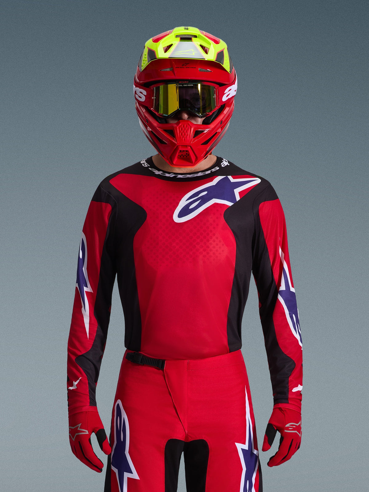 Maillot Alpinestars Fluid Grid 2026, maillot de motocross, rouge, noir et violet, porté par un pilote avec un casque rouge et jaune fluo et des lunettes dorées, avec de grands logos étoilés sur la poitrine et les manches, équipement coordonné pour la course tout-terrain