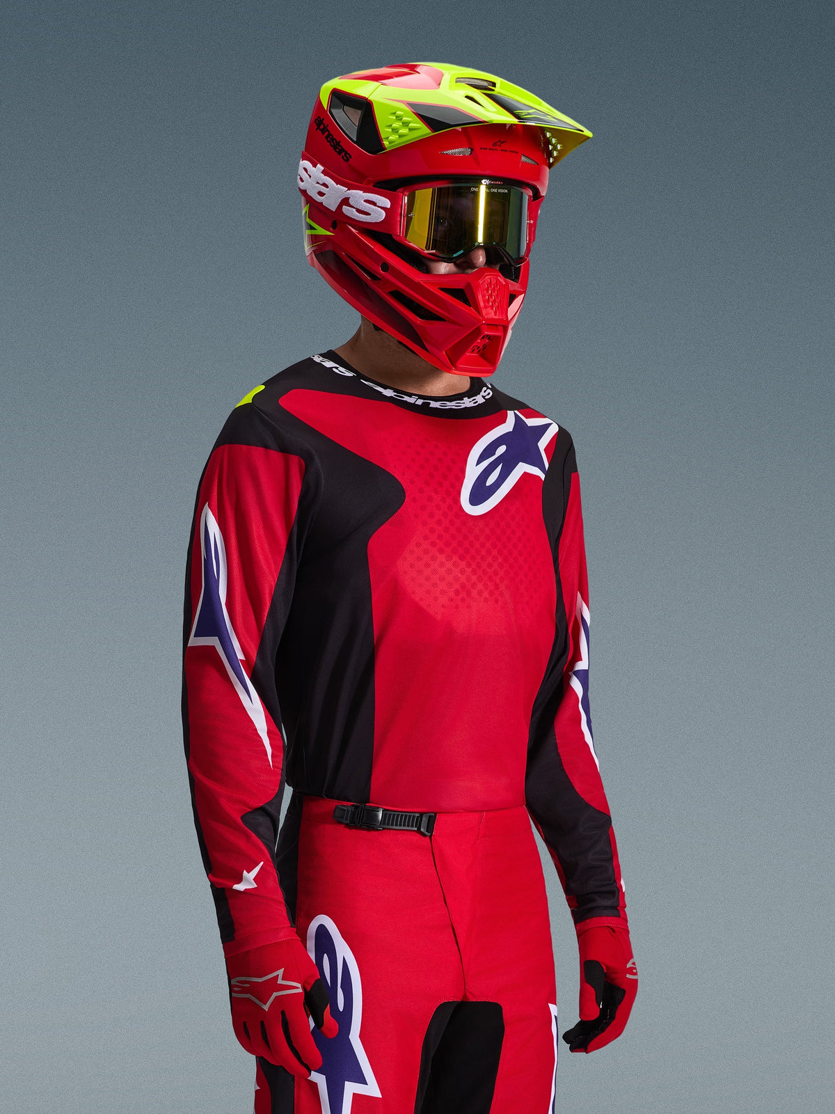 Pilote de motocross portant le maillot Alpinestars 2026 Fluid Grid rouge, noir et violet, avec un pantalon de motocross assorti, un casque rouge aux accents jaune fluo, des lunettes dorées et des gants rouges, tenue de course tout-terrain complète