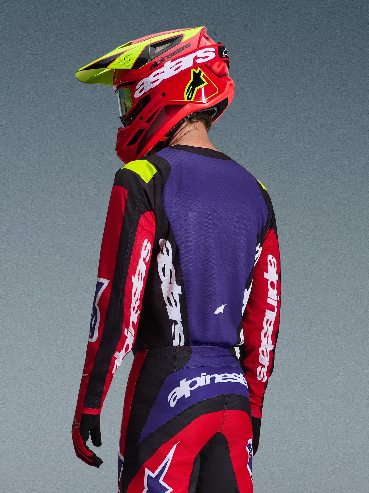 Pilote de motocross portant le maillot Alpinestars 2026 Fluid Grid, rouge, noir et violet, vue de dos montrant le panneau arrière violet et les manches rouges avec logos blancs, pantalon assorti et casque rouge et jaune fluo, tenue de course coordonnée