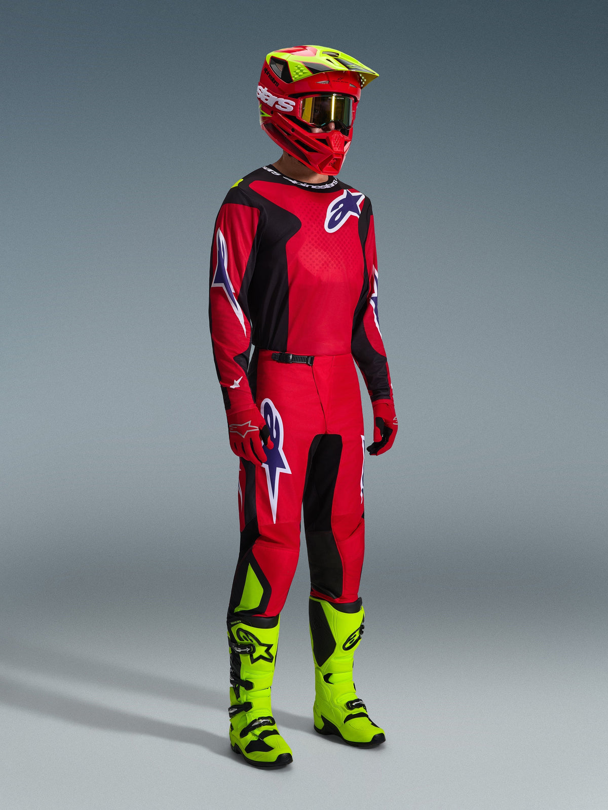 Pilote de motocross en tenue de course complète Alpinestars, maillot Fluid Grid 2026 rouge, noir et violet, pantalon assorti avec accents de logo violets, casque rouge et jaune fluo avec lunettes dorées, et bottes jaune fluo, tenue coordonnée pour le tout-terrain
