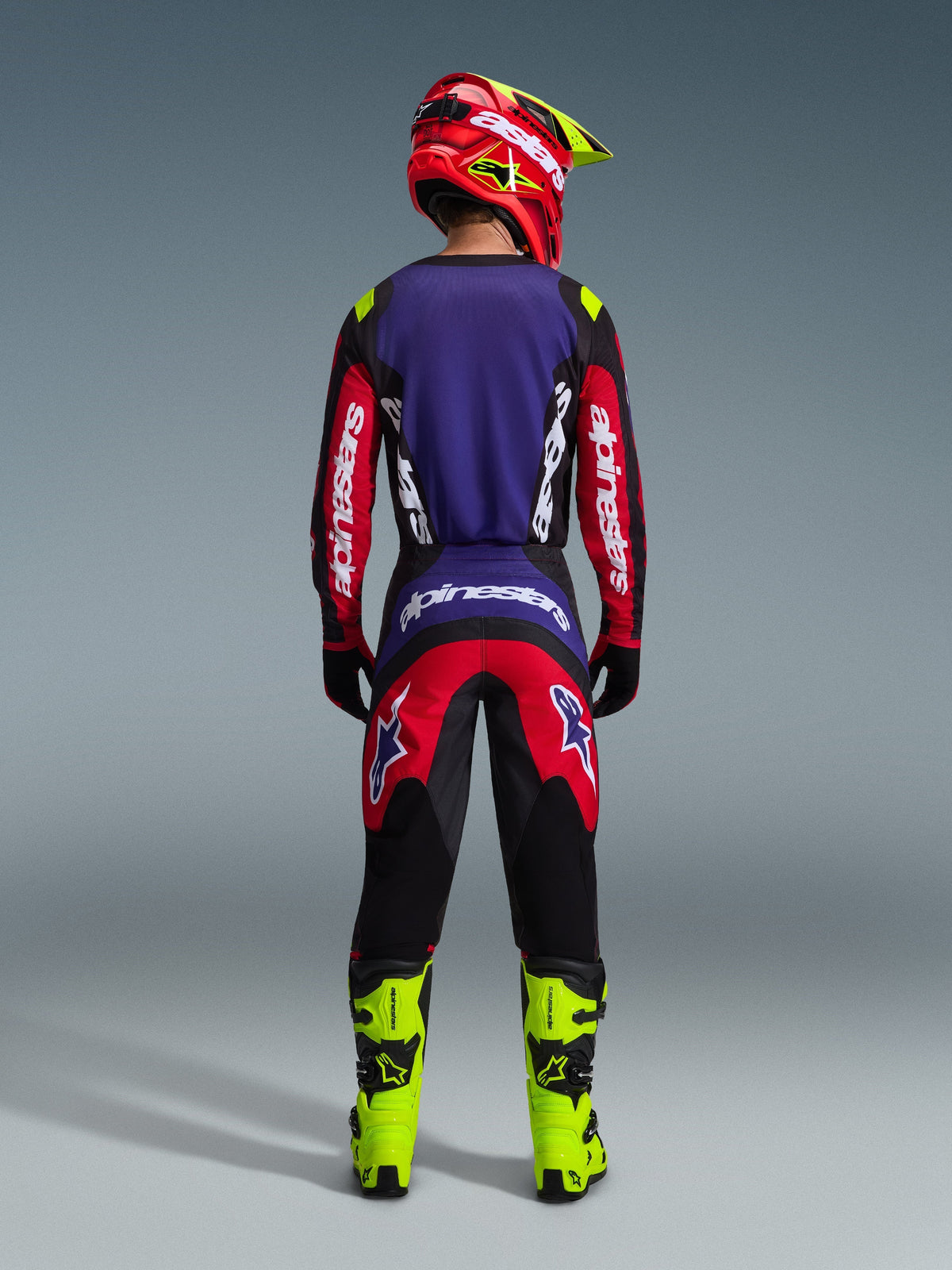 Pilote de motocross vu de dos portant le maillot Alpinestars Fluid Grid 2026 en Rouge Noir Violet, avec logos Alpinestars blancs sur les manches longues, pantalon de motocross assorti, bottes jaune fluo et casque rouge et jaune, tenue complète pour la course tout-terrain