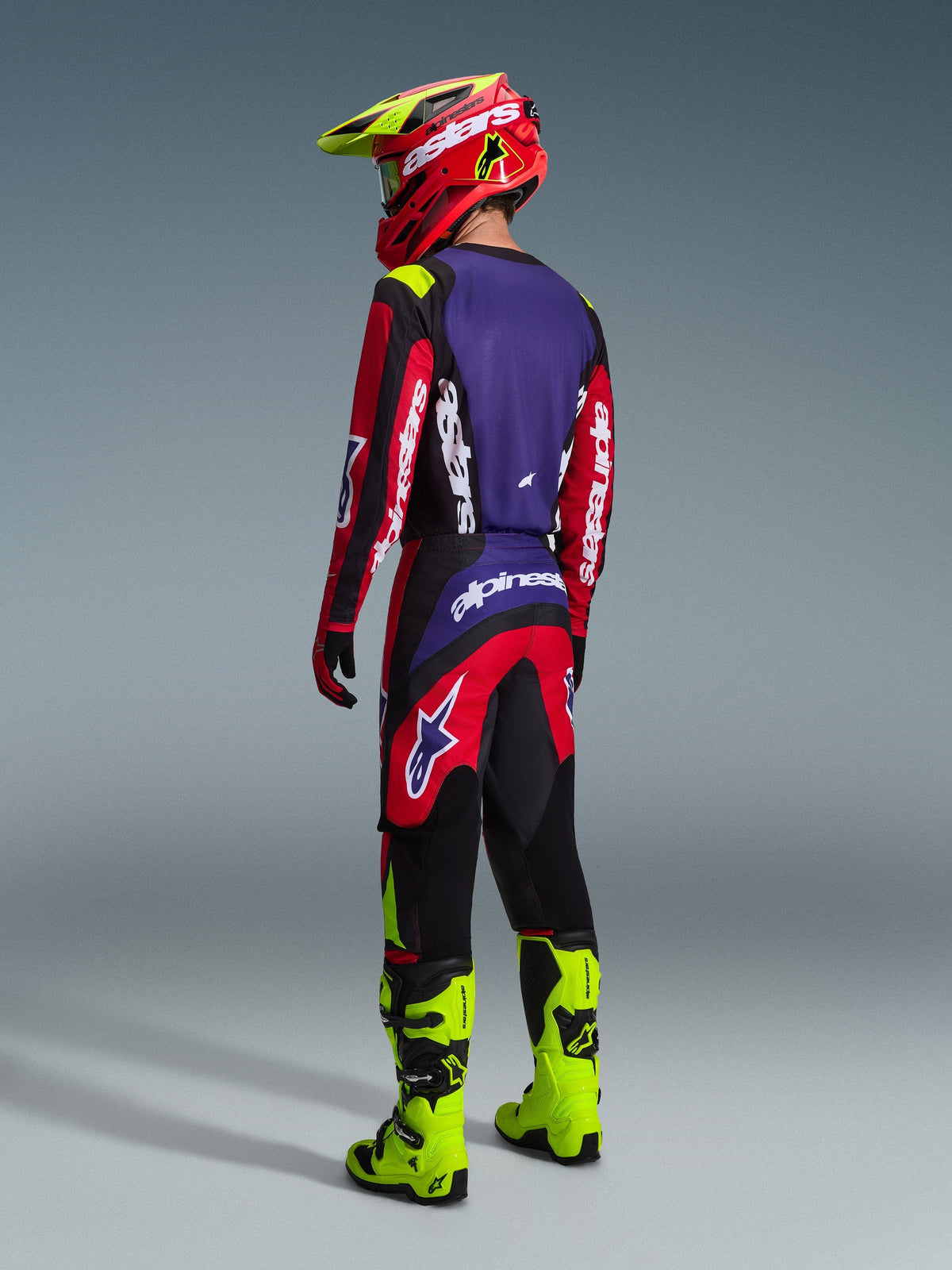 Pilote de motocross vu de dos, portant le maillot Alpinestars 2026 Fluid Grid et le pantalon assorti en rouge, noir et violet, avec des logos blancs, associé à des bottes jaune fluo et un casque rouge et jaune, tenue de course tout-terrain complète