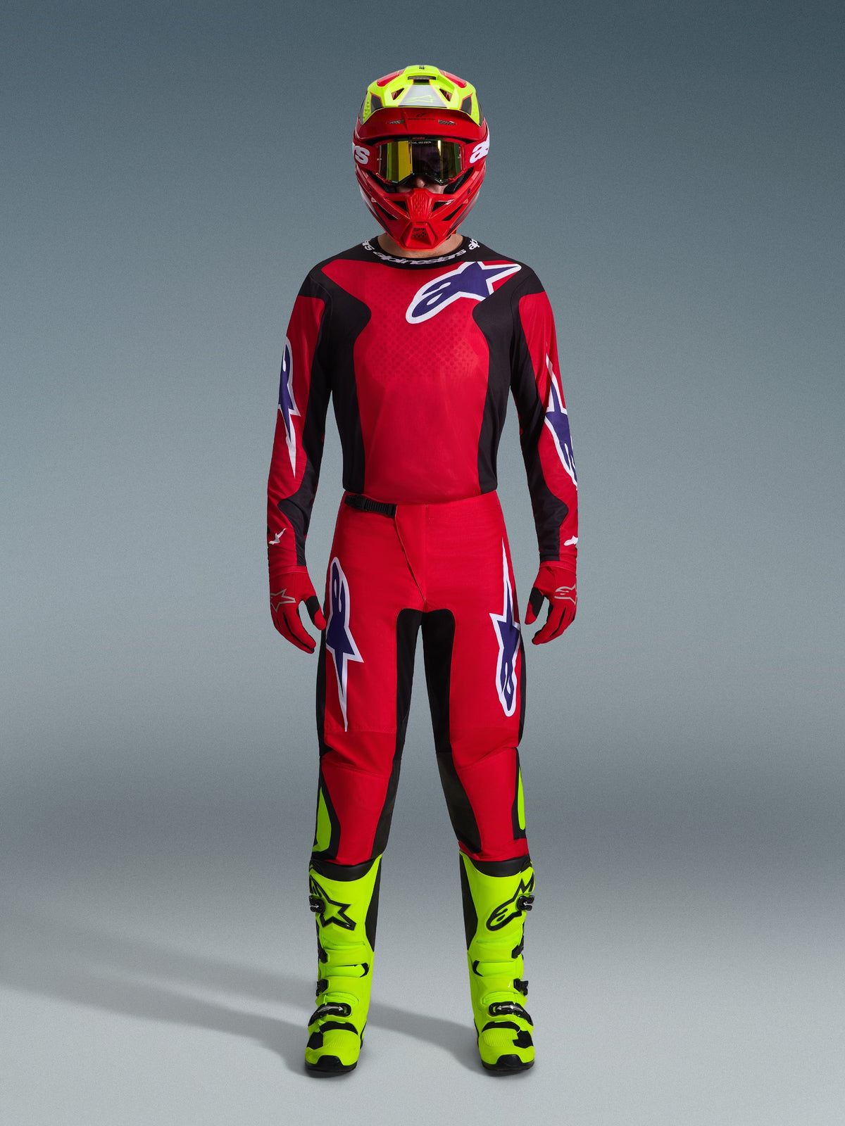 Pilote de motocross portant le maillot Fluid Grid 2026 en rouge, noir et violet, pantalon rouge assorti avec logos violets, casque rouge avec accents jaune fluo, bottes jaune fluo, tenue de course complète coordonnée pour le motocross tout-terrain