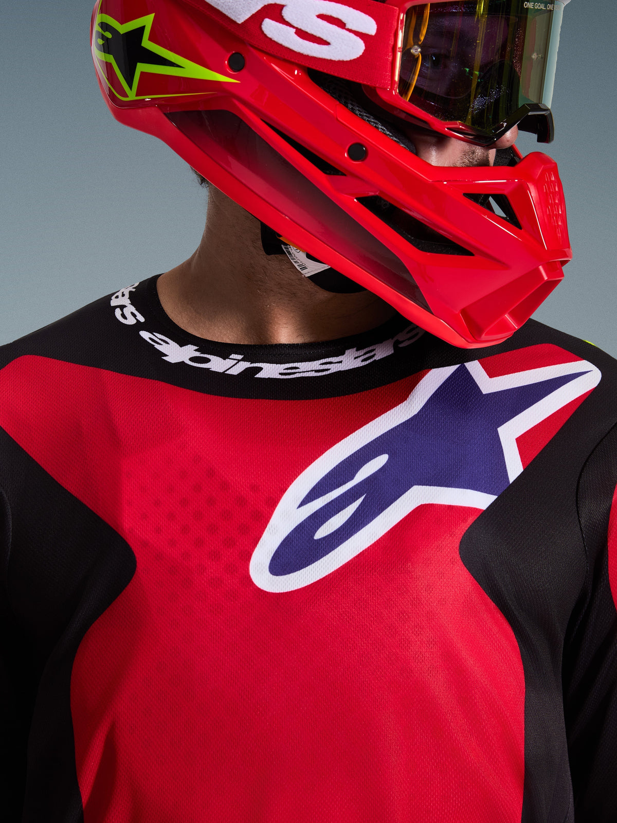Gros plan d'un pilote de motocross portant le maillot Alpinestars 2026 Fluid Grid en rouge, noir et violet, avec un grand logo sur la poitrine et un col sport griffé, associé à un casque rouge et des lunettes, mettant en avant le poly-tissu technique et la coupe athlétique.