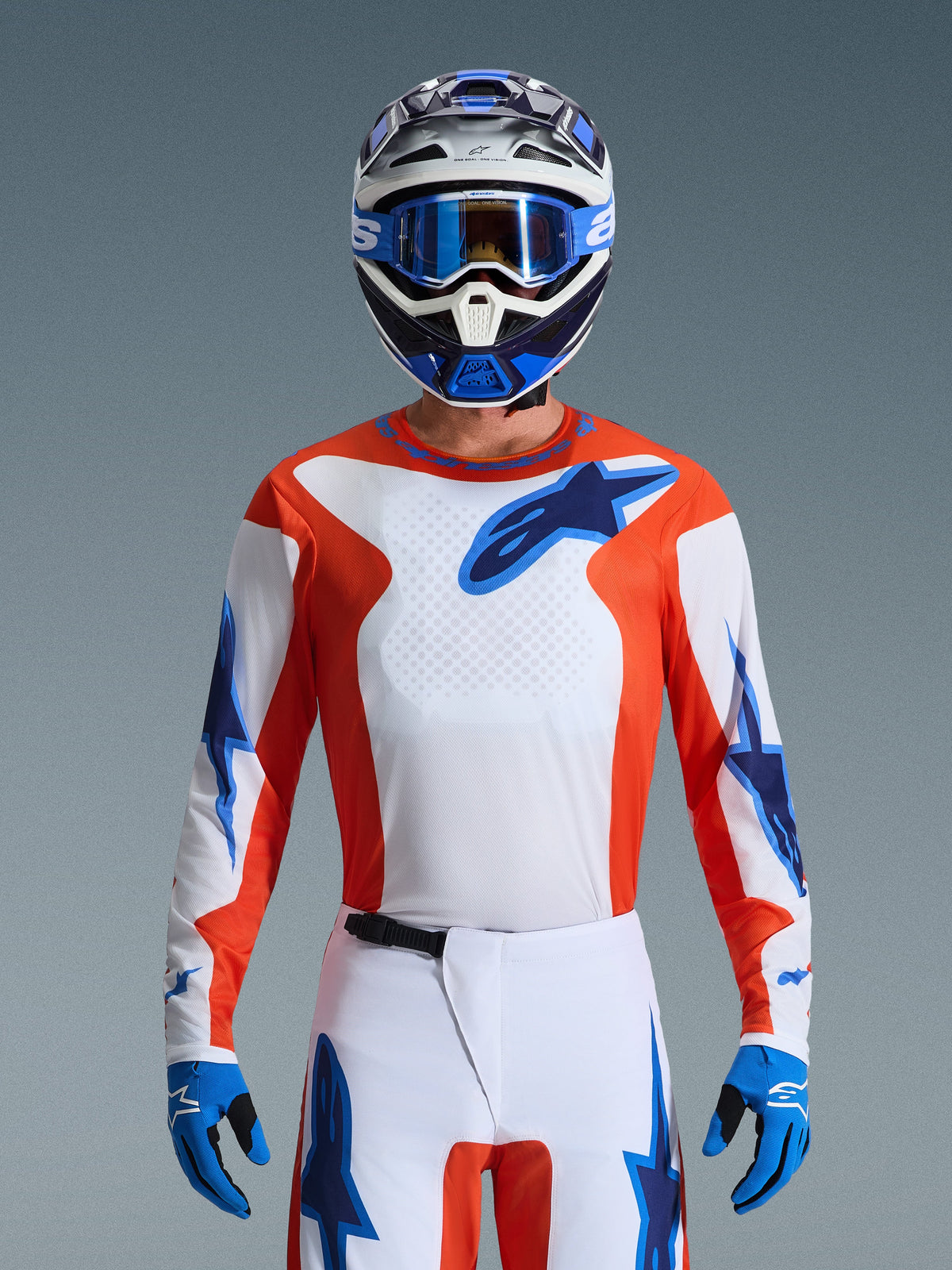 Ensemble d'équipement Alpinestars 2026 Fluid Grid, pilote de motocross portant un maillot orange et blanc avec des logos d'étoiles bleus, pantalon blanc avec accents bleus, casque bleu et blanc assorti avec lunettes et gants bleus, tenue de course tout-terrain complète