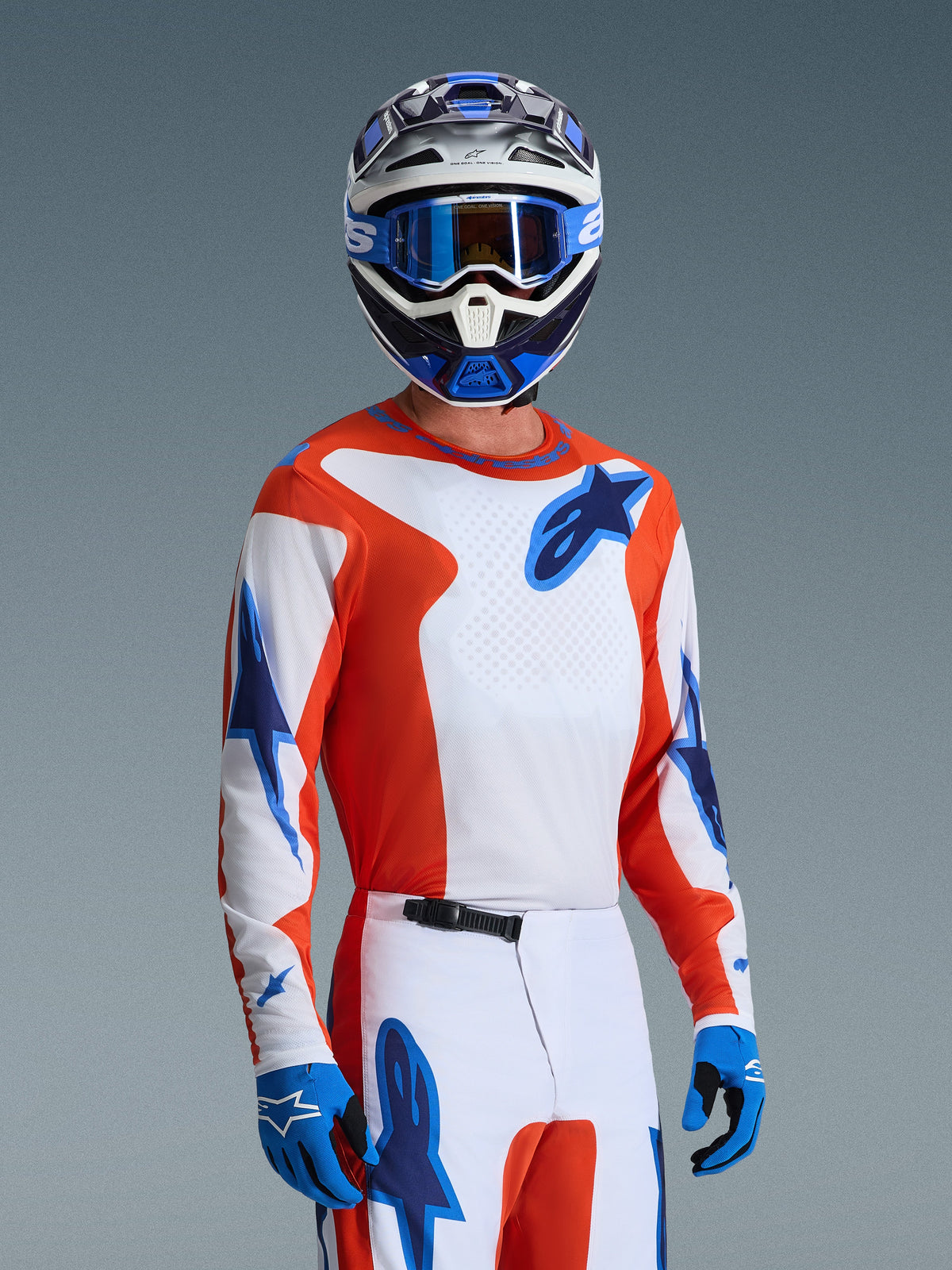 Pilote de motocross portant l'ensemble d'équipement Alpinestars Fluid Grid 2026, maillot orange et blanc avec logos d'étoiles bleus, pantalon blanc et orange avec accents bleus, gants bleus, casque bleu et blanc avec lunettes bleues, tenue de course athlétique coordonnée pour la performance tout-terrain