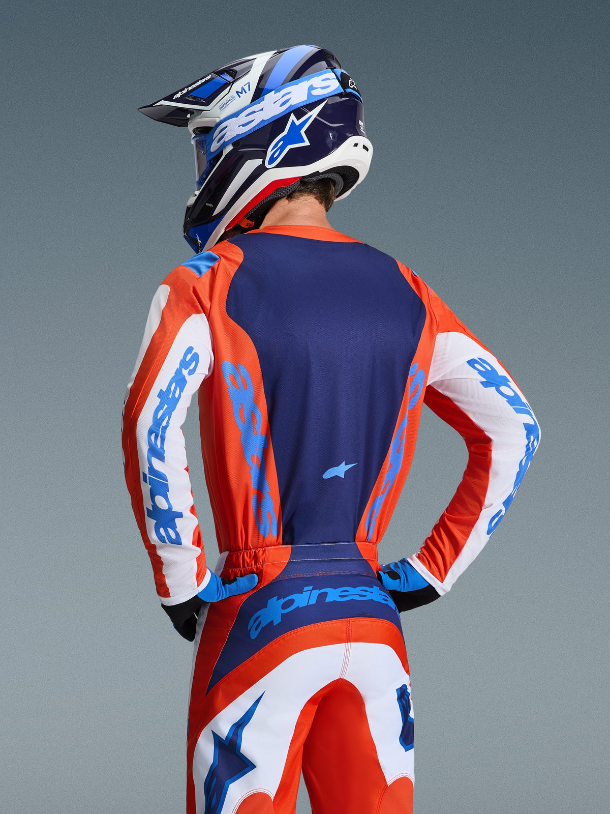 Maillot Alpinestars Fluid Grid 2026, vue arrière d'un pilote de motocross portant une tenue coordonnée orange et bleu UCLA, panneau arrière bleu marine, accents blancs sur les manches avec marquage bleu, casque assorti, équipement tout-terrain haute performance