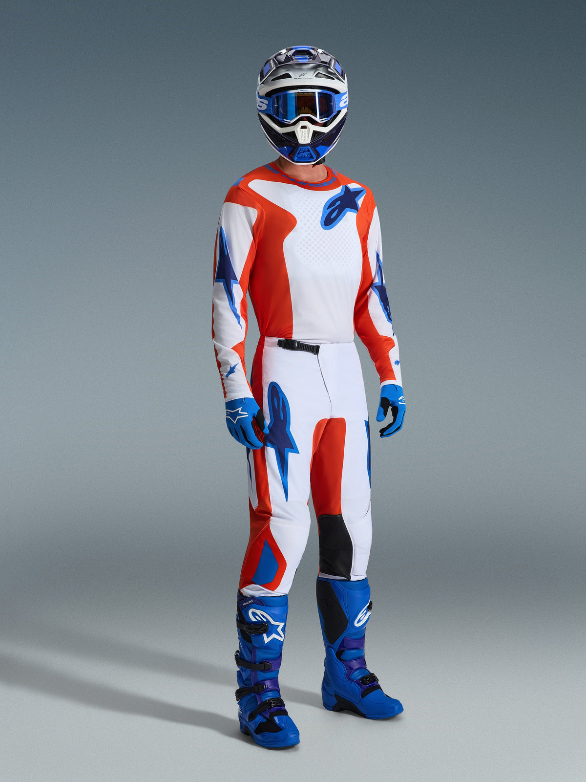Pilote de motocross portant l'ensemble d'équipement Alpinestars Fluid Grid 2026, maillot et pantalon blancs avec des graphismes orange et bleu UCLA, casque et lunettes bleus assortis, gants bleus et bottes de course bleu vif, tenue de performance coordonnée pour la conduite tout-terrain.