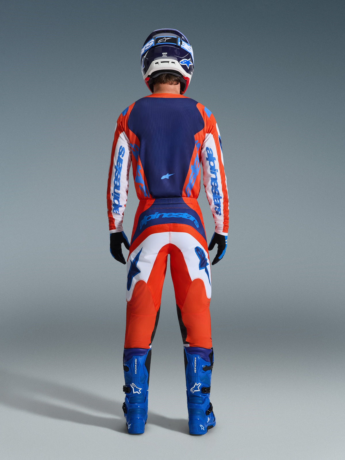 Maillot Alpinestars Fluid Grid 2026, maillot de motocross, orange et bleu UCLA, vue arrière d'un pilote en tenue de course complète, comprenant un panneau arrière bleu foncé et des manches blanches avec logos bleus, associé à un pantalon assorti et des bottes bleues.