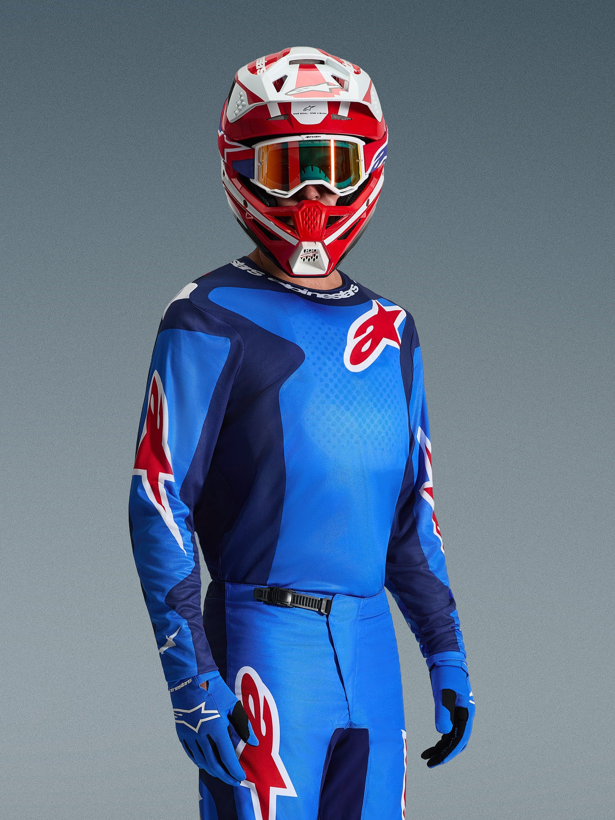Pilote de motocross portant le maillot Alpinestars Fluid Grid 2026 en bleu et rouge, avec des logos étoiles Alpinestars rouges, associé à un pantalon bleu assorti, des gants bleus et un casque rouge et blanc avec lunettes
