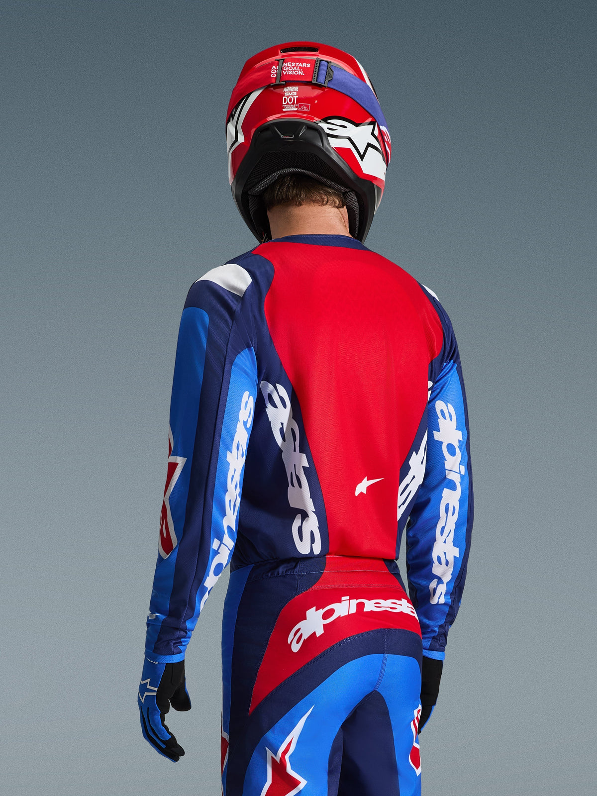 Vue arrière d'un pilote de motocross portant le maillot Alpinestars Fluid Grid 2026 en bleu et rouge, avec des logos blancs sur les manches et le dos, associé à un pantalon de motocross assorti et un casque rouge pour une tenue de course tout-terrain complète