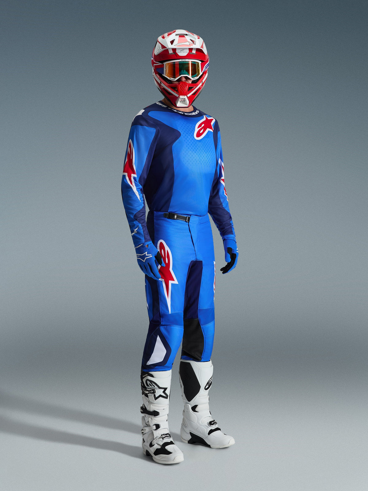 Pilote de motocross portant le maillot Alpinestars 2026 Fluid Grid en Bleu Rouge, avec pantalon de motocross assorti, bottes blanches et noires, et casque rouge et blanc, ensemble d'équipement professionnel pour la performance tout-terrain