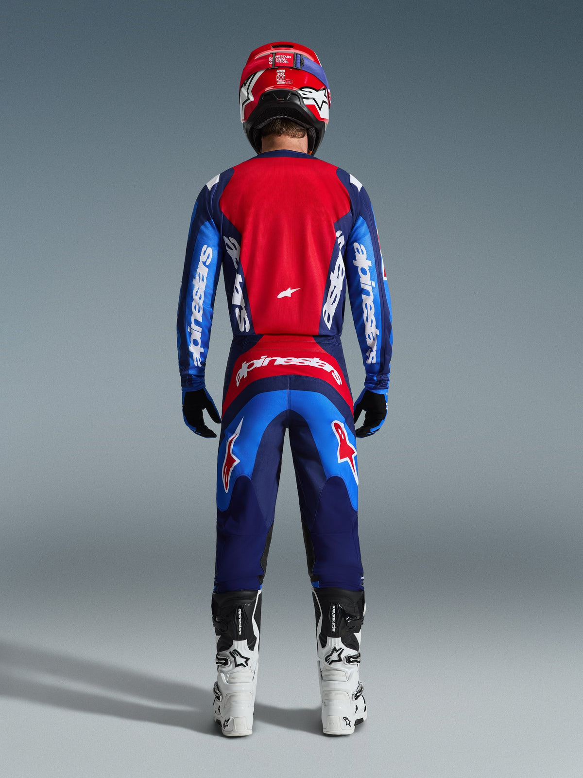 Vue arrière d'un pilote de motocross portant le maillot Alpinestars Fluid Grid 2026 bleu et rouge, doté d'un large panneau dorsal rouge et de manches bleues avec marquage Alpinestars blanc, associé à un pantalon assorti, des bottes blanches et noires et un casque rouge pour une tenue de course tout-terrain complète.