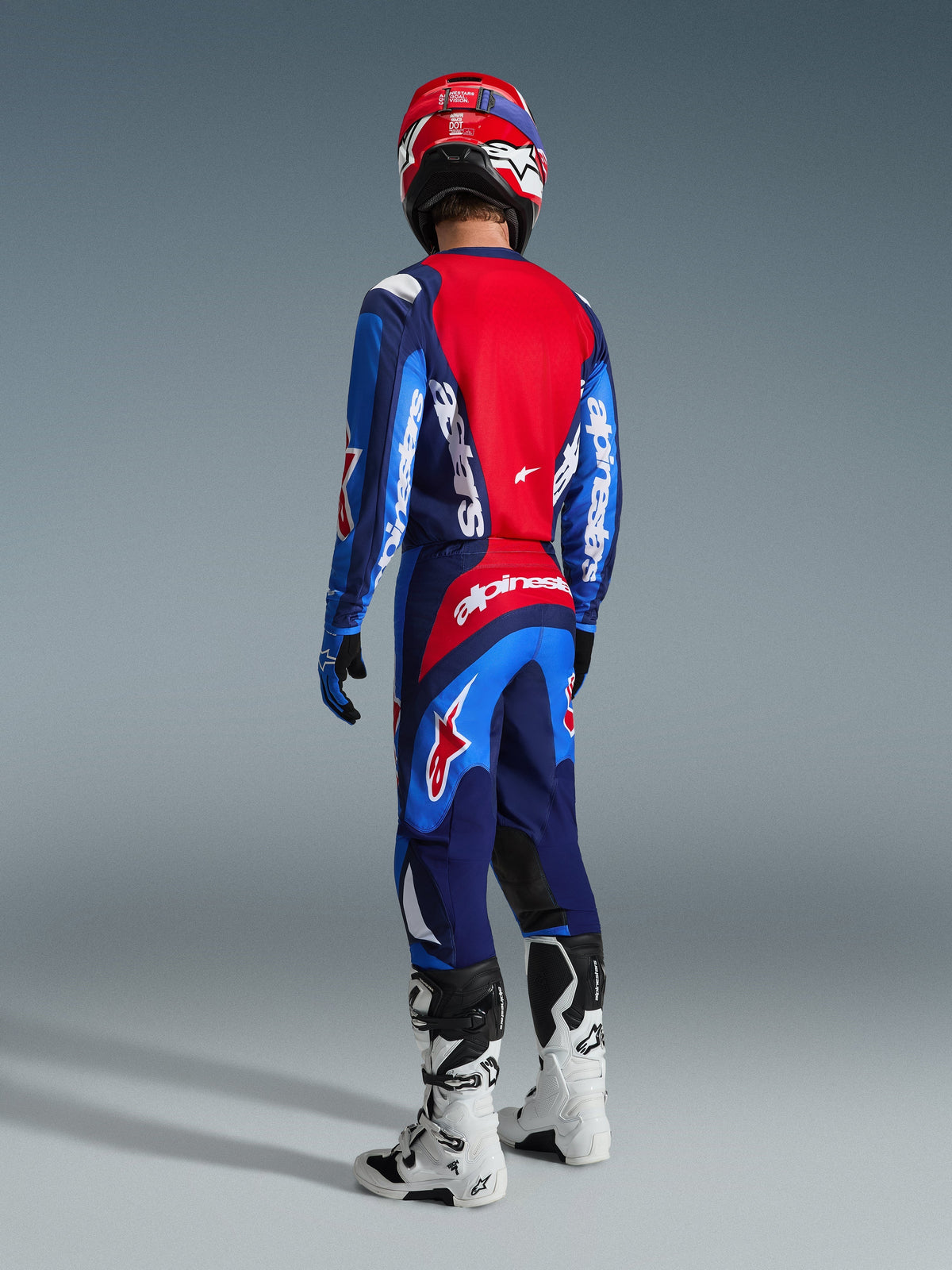 Pilote de motocross vu de dos portant le maillot Alpinestars 2026 Fluid Grid bleu et rouge, pantalon de motocross assorti avec un marquage blanc imposant, casque rouge et bottes blanches et noires, tenue de course tout-terrain complète