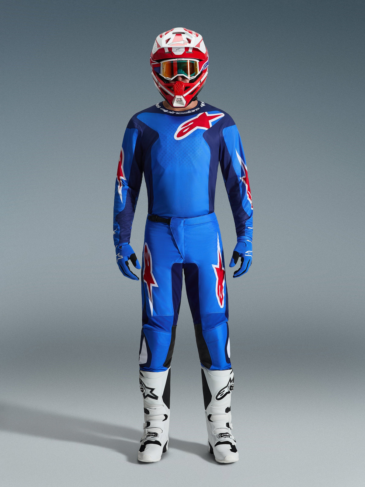 Pilote de motocross portant le maillot Alpinestars 2026 Fluid Grid bleu et rouge, pantalon bleu assorti avec logos d'étoiles blanches et rouges, casque rouge et blanc avec lunettes miroir, gants bleus et bottes de course blanches et noires, ensemble complet d'équipement tout-terrain